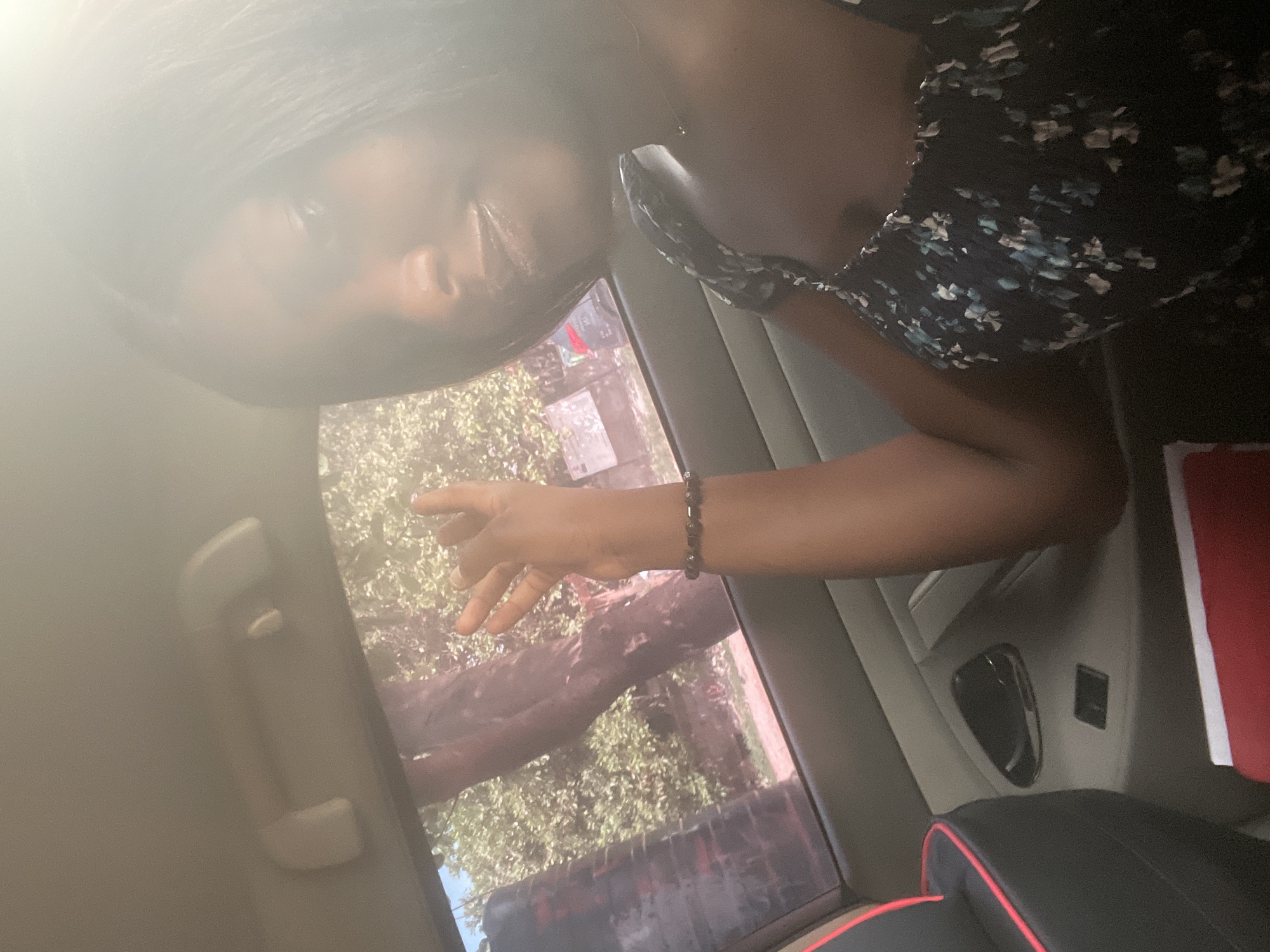 An overhead, close-up shot taken inside a car, likely in motion. A dark-skinned woman with long, dark hair is partially visible at the top of the frame. She is wearing a floral print dress. Her arm, adorned with a beaded bracelet, extends towards the car window. Through the window, trees and a building are visible, suggesting an outdoor setting, likely in Kurunduma, Nigeria. The car's interior includes a grab handle, side panel, and a black and red headrest. A partially visible red object is present.