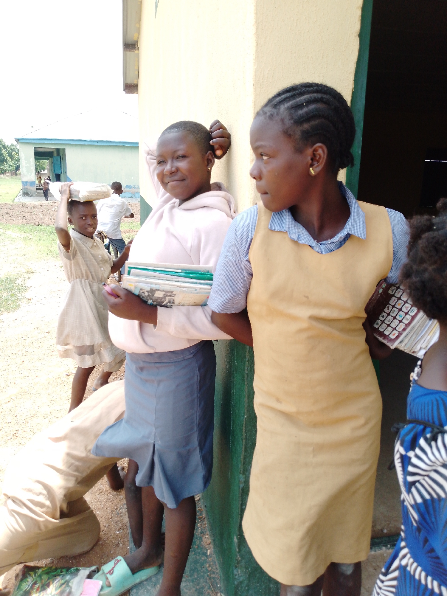In Akwanga, Nigeria, the image shows two schoolgirls standing near a building with a pale yellow exterior and green trim. The girl on the left wears a light pink sweatshirt and a grey skirt. She has books in her arms and leans against the wall. The girl on the right wears a blue and white striped shirt with a tan jumper and carries books. Another young girl is visible in the background carrying a white bag on her head. A man is walking behind her in the distance. Other people are standing nearby. A sandy area is in the foreground, and there is a building in the background.