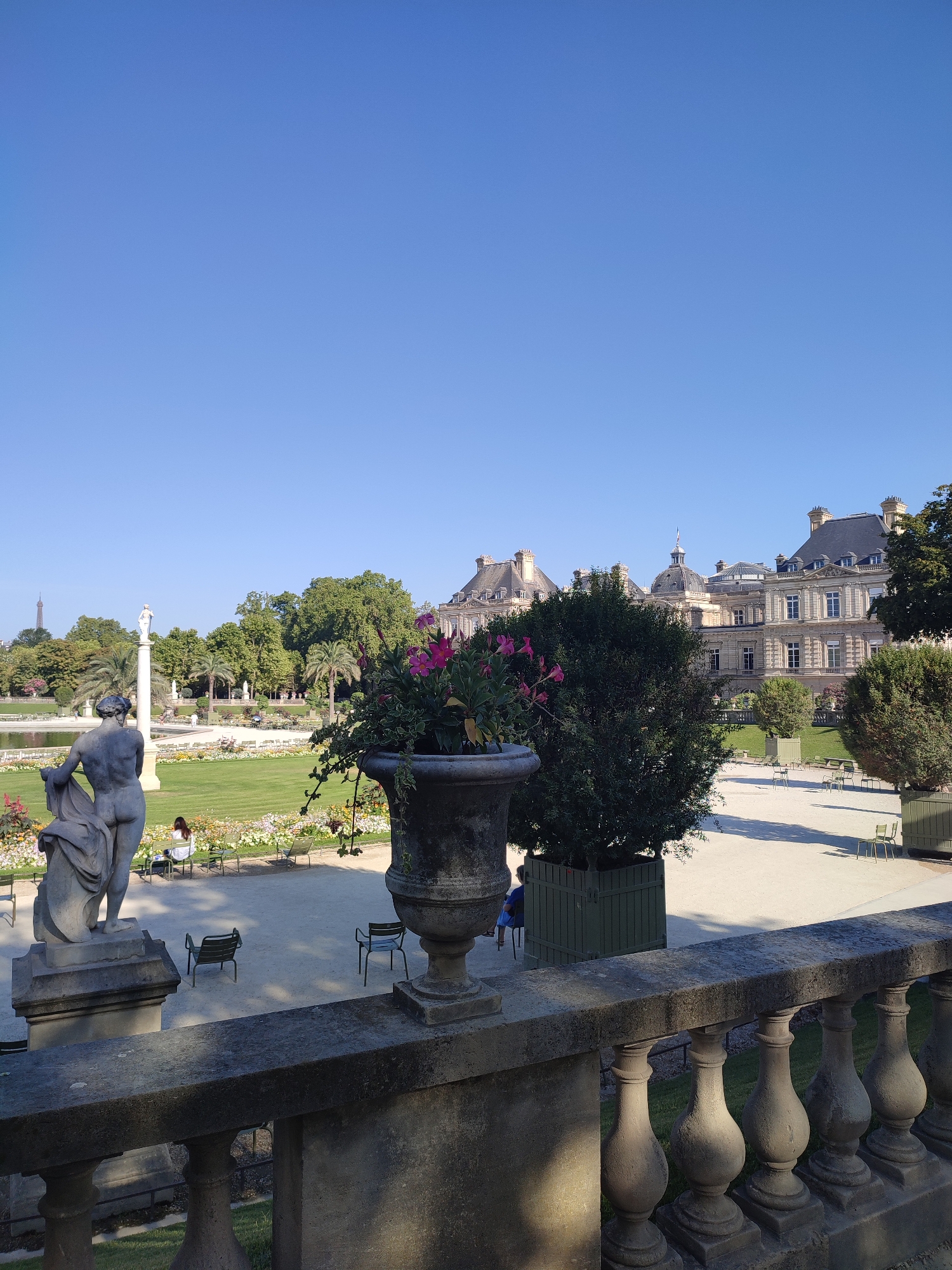 The image captures a view of the Luxembourg Garden in Paris, France. In the foreground, a stone balustrade with ornate pillars runs horizontally across the bottom of the frame. To the left, a partial view of a classical stone sculpture of a woman is visible, positioned on a pedestal. Beyond this, a large, ornate stone planter overflows with vibrant pink flowers and trailing greenery.
The middle ground of the image features a neatly manicured lawn dotted with flowerbeds and interspersed with green metal chairs. A few individuals are seated on these chairs, with one person in the left-middle ground facing away from the camera. Further back, a tranquil pond reflects the clear blue sky. To the right of the pond, a stately building with classical architecture, featuring multiple windows, dormers, and domes, stands prominently. Green trees and palm trees are scattered throughout the garden, with the Eiffel Tower visible in the far left background. The overall scene depicts a peaceful, sunny day in a Parisian park.