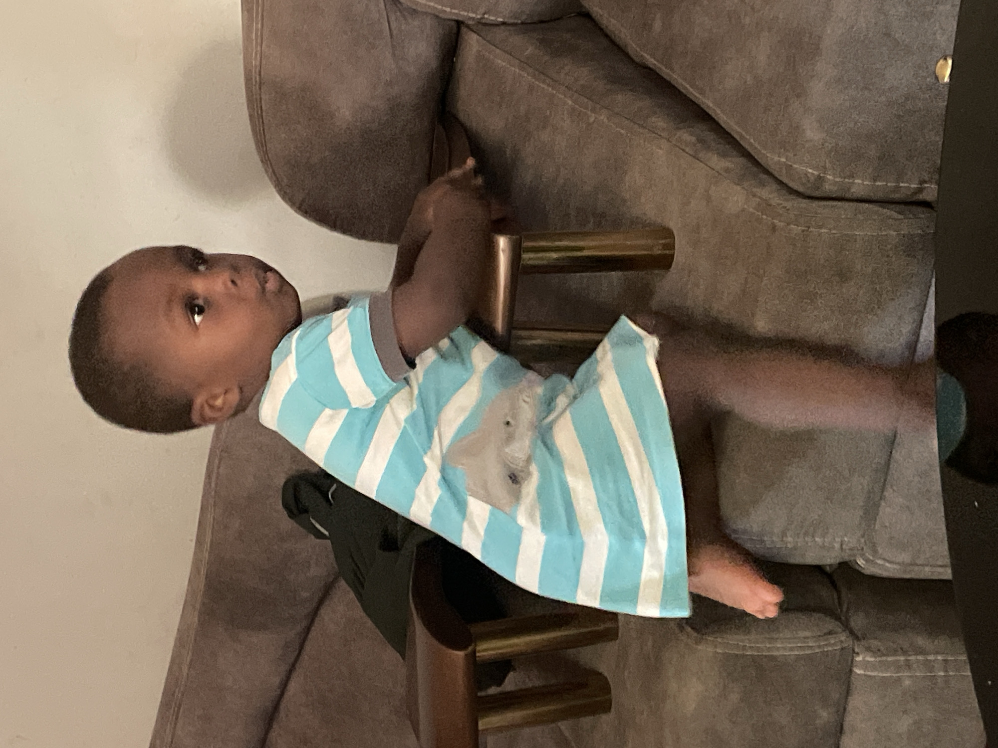 A young child is lying on a gray upholstered sofa, wearing a light blue and white striped shirt with a light gray elephant design on the front. The child is positioned near a small, dark brown wooden table. The background is a light beige wall. The image was taken indoors. It is not possible to identify the exact location in Jos, Nigeria from the information provided.