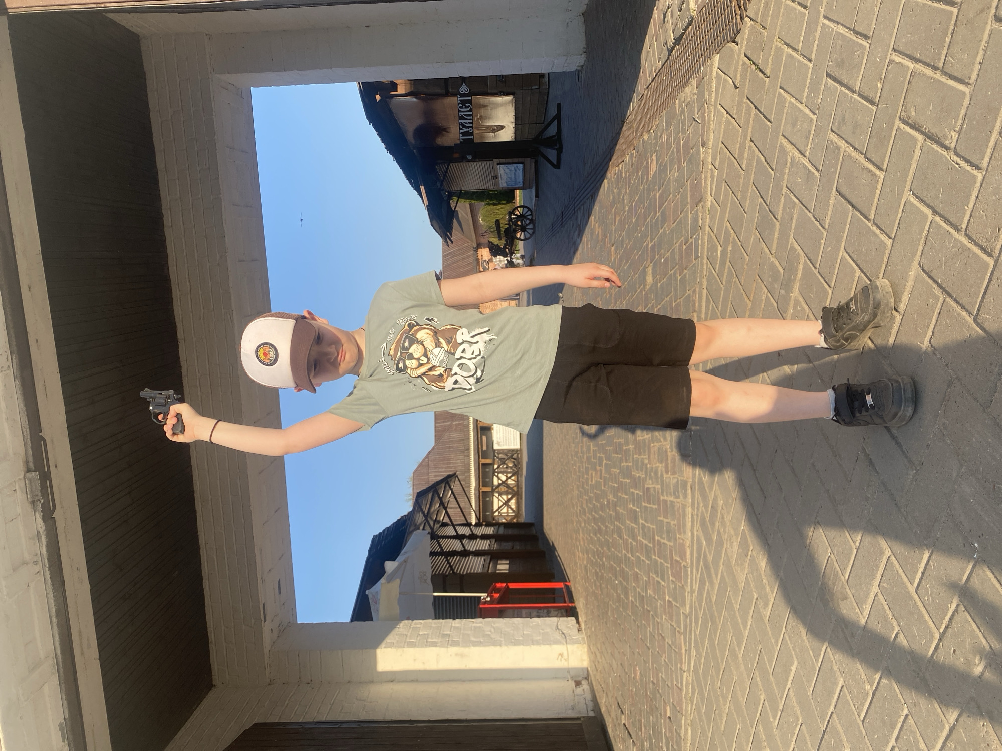 A child is standing on a brick path, holding a handgun. The backdrop features buildings, including an older, wooden structure with a sign in Russian that seems to say "Toilet". This points to a location in Sviyazhsk, Russia, a town known for its historical architecture. The child is wearing a cap, t-shirt, and shorts, and they are positioned within a stone entryway.