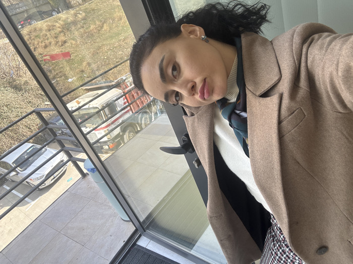 A woman with dark, curly hair pulled back poses for a selfie, looking directly at the camera with a slight smile. She is wearing a light brown double-breasted coat over a white turtleneck sweater and a patterned scarf, with a red, black, and white houndstooth skirt visible below. The scene is captured from indoors, looking out through a large glass panel.
Outside the glass, a light-tiled patio with a black metal railing is visible. Beyond the railing, a parking area features a white Ford Fusion sedan and a white and green truck with a red stripe. The truck has "თბილისის მერია" (Tbilisi City Hall) inscribed on its side, identifying the location as Tbilisi, Georgia. A light blue waste bin stands near the railing. Further in the background, a dry, grassy slope with sparse vegetation and dirt is visible, with other vehicles parked on an elevated surface beyond it. The scene is illuminated by bright sunlight, causing some reflections on the glass.