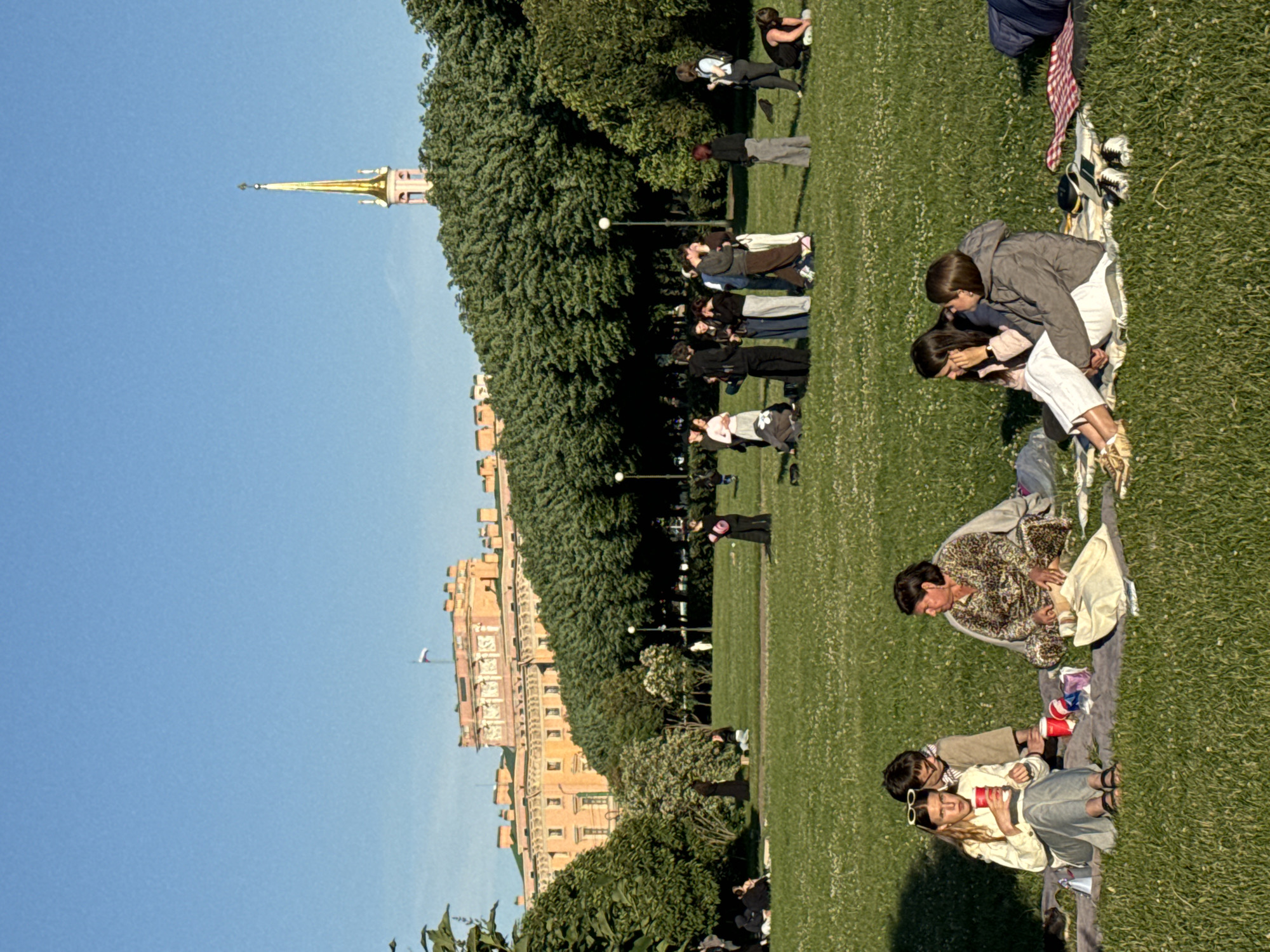 A group of people are picnicking on the grass in a park in Saint Petersburg, Russia. In the background, there is a large, ornate building with a golden spire, likely a palace or museum, and a dense line of trees. The sky is clear and blue. The people are scattered across the lawn, some sitting on blankets, others standing in small groups. Some are holding drinks, and there are picnic items visible on the blankets. The overall atmosphere is relaxed and leisurely, suggesting a warm, sunny day. The foreground shows a few individuals sitting closely together, engaged in conversation or observing their surroundings. The midground has several groups of people, some interacting closely, while others are more spread out. The background reveals the architectural grandeur of the city.