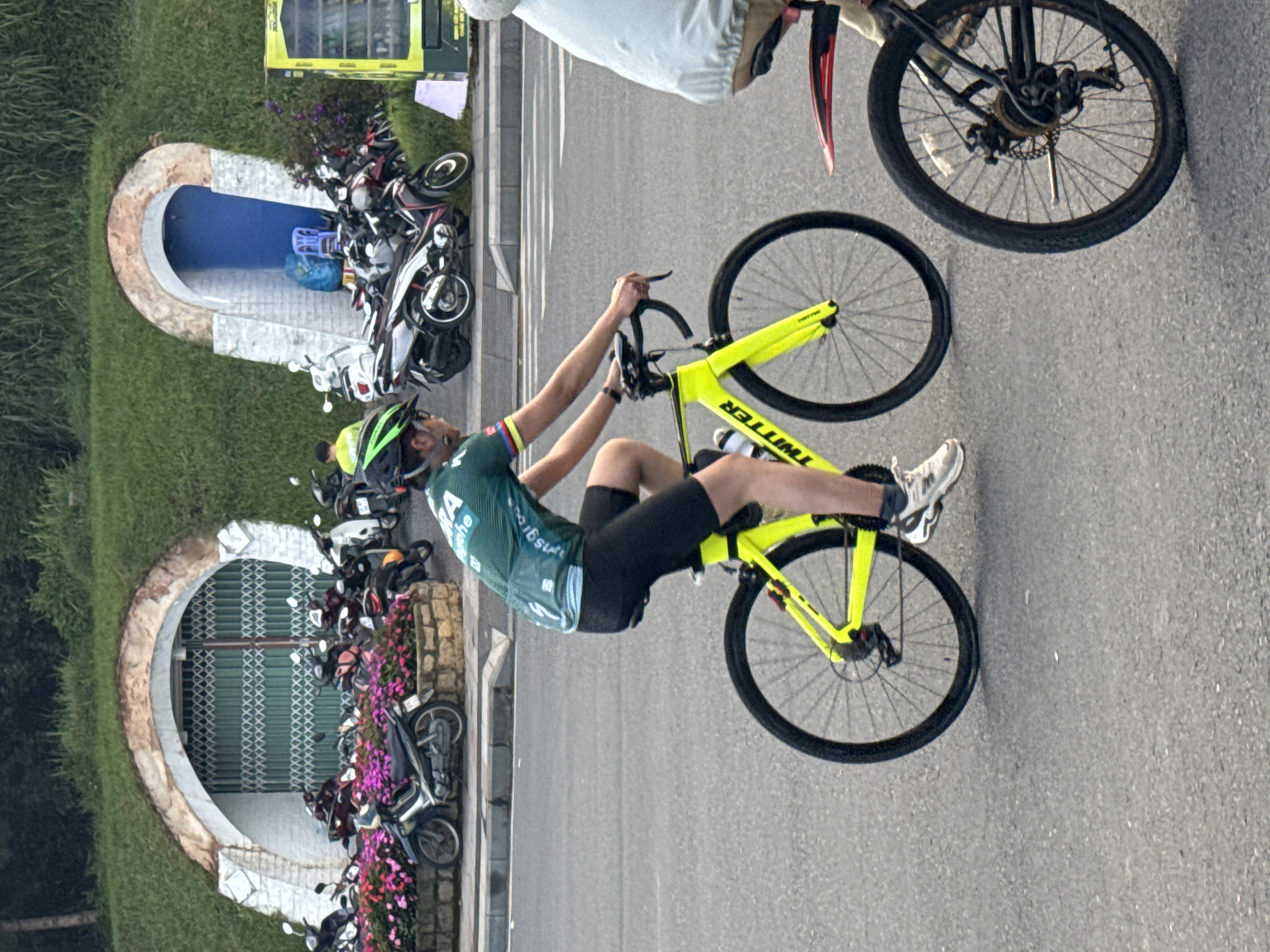 A cyclist in a green and teal jersey and black cycling shorts rides a bright yellow road bike with "TWITTER" printed on the frame. The cyclist wears a green helmet and white athletic shoes. The scene appears to be a street in Da Lat, Vietnam, with a grassy embankment featuring two tunnel-like structures in the background. The left tunnel entrance is covered by a green, collapsible metal gate, and the right entrance has a blue door. Several parked motorcycles are visible near the tunnels, alongside flower beds and a vending machine. The road is paved asphalt.