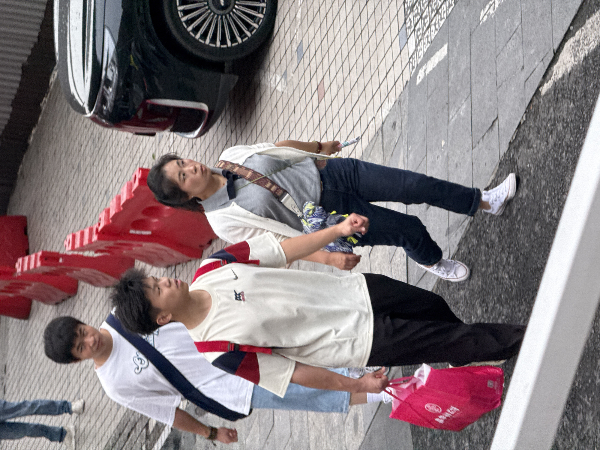 In Yuzhong District, China, three young adults walk on a tiled sidewalk adjacent to a street. A person in the foreground, wearing a white t-shirt with red and blue accents and black pants, carries a bright pink shopping bag. To their left, a second individual in a white t-shirt with a navy blue strap walks in the same direction. Behind them, a third person, a woman wearing a grey polo shirt and a white cardigan with dark blue jeans, walks with a crossbody bag. A dark-colored vehicle is parked to the right of the scene. Red barriers are visible in the background, suggesting a construction or event area.