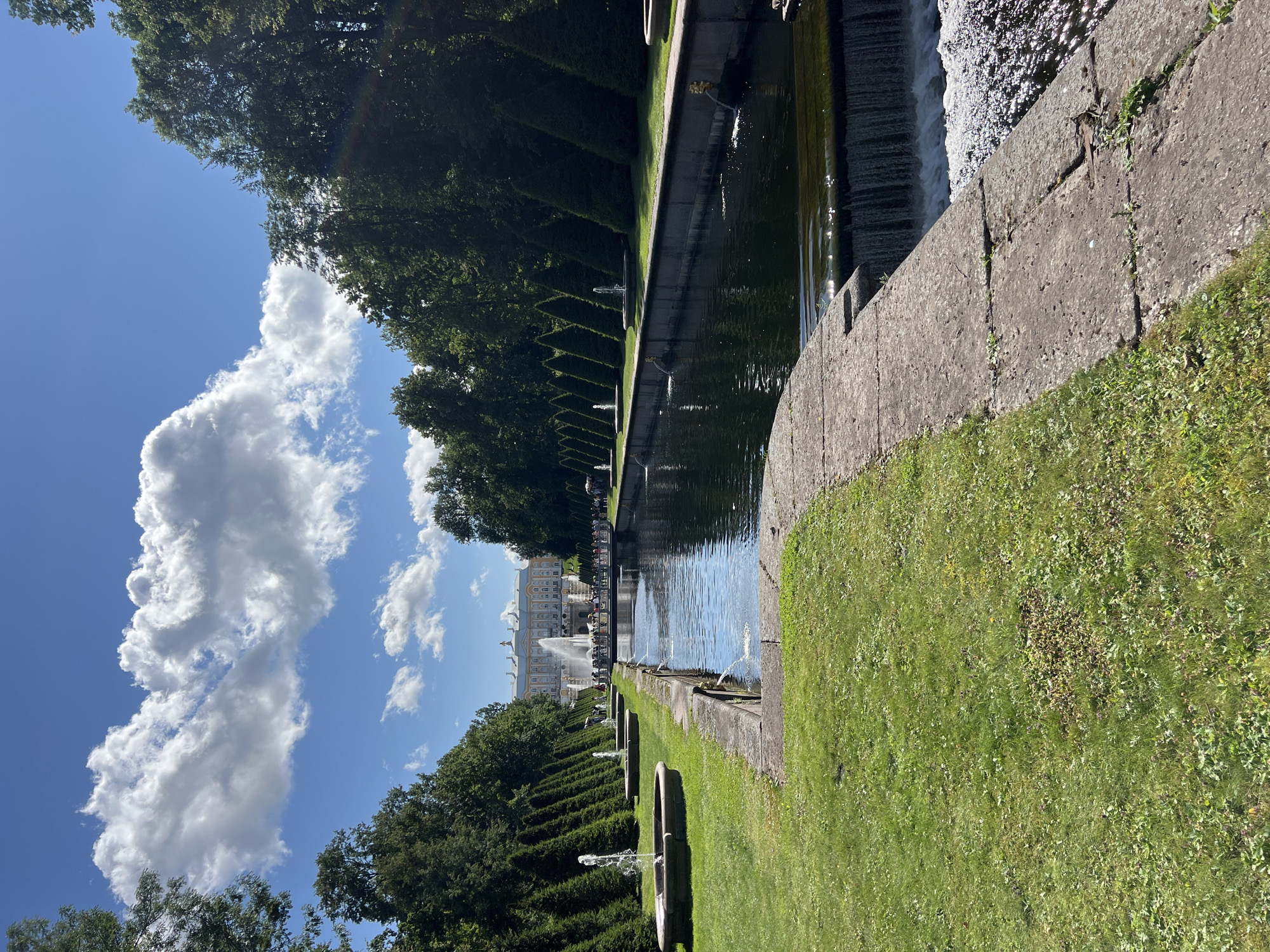 This image depicts a view of the Grand Cascade and Samson Fountain at Peterhof Palace in Peterhof, Russia. The scene is dominated by a formal garden with manicured lawns, rows of conical trees, and a long rectangular canal. Water from multiple fountains is visible, including individual smaller fountains along the canal and a large, multi-tiered fountain in the distance in front of a grand palace. A crowd of people is gathered on a bridge in front of the palace. The sky is blue with scattered white clouds. The foreground shows a stone pathway bordering a grassy area with a small waterfall cascading into the canal.
