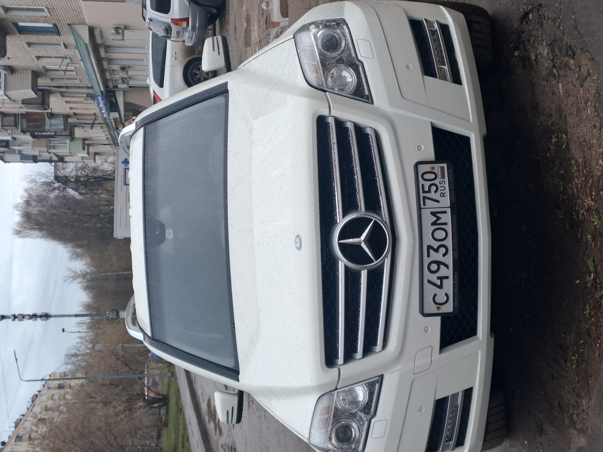 A white Mercedes-Benz SUV is parked outdoors on a street with rain drops visible on its surface. The front of the vehicle is facing the viewer, prominently displaying the grill with the Mercedes-Benz logo and a Russian license plate, C493OM 750 RUS, with the text "МБ-ИЗМАЙЛОВО" and "(495) 66-00000". In the background, other cars, buildings and trees can be seen under an overcast sky. The location is in the city of Elektrostal, Russia.