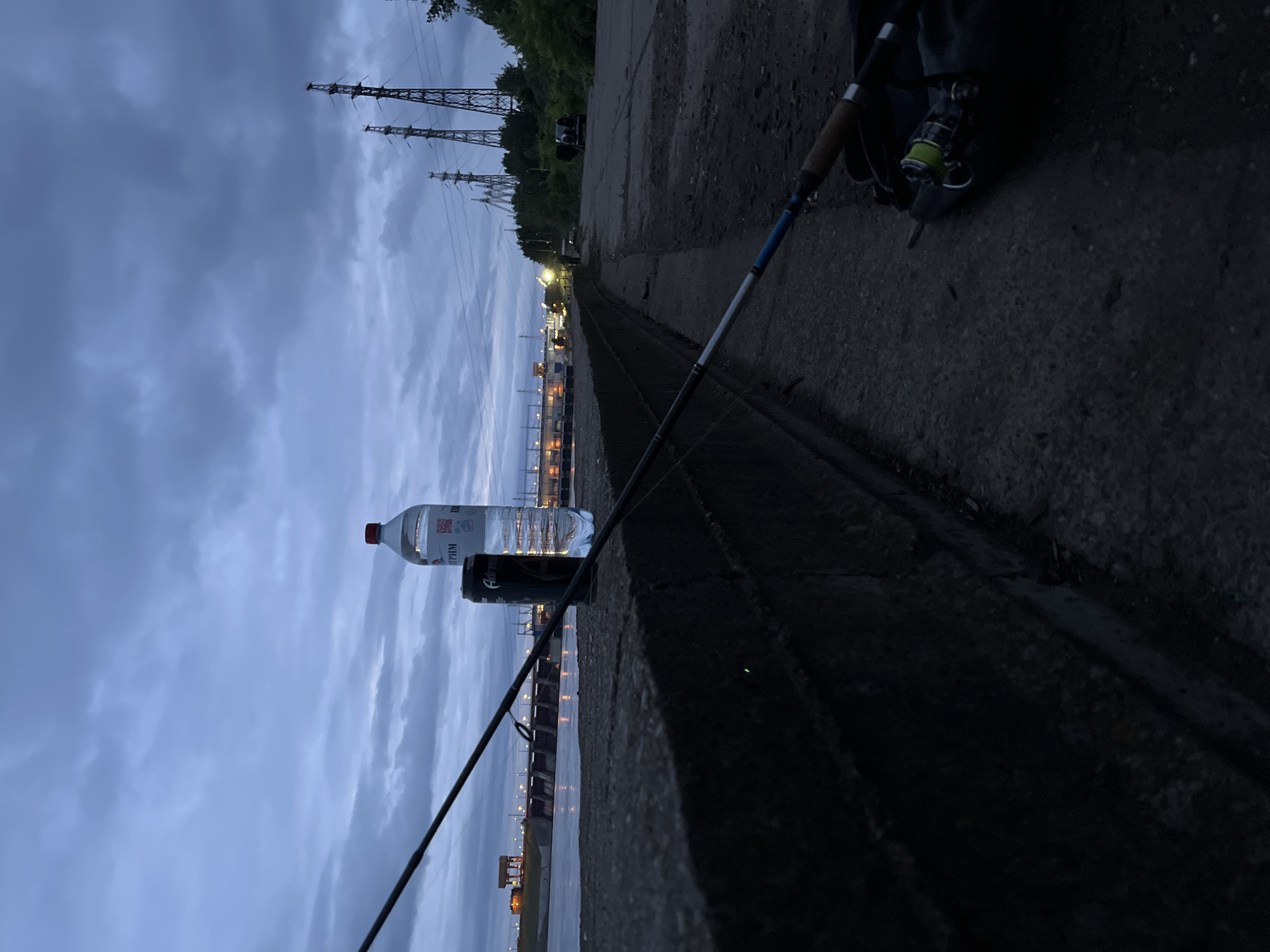 This image, taken at dusk or dawn in Noviy, Russia, captures a fishing setup along a concrete embankment beside a large body of water. In the foreground, a fishing rod with a yellow-green line reel, resting on a black bag, lies on a dark concrete path. On the adjacent concrete embankment, a clear plastic water bottle with a red cap and a "GAZPROM" label stands next to a black "Adrenaline" energy drink can. Across the water, a large, illuminated dam or hydroelectric power station is visible, with its lights reflecting on the water's surface. In the background, multiple tall electricity pylons with power lines traverse the cloudy, dark blue sky. Further along the path on the right, a dark vehicle with an open rear door is parked near trees. No individuals are clearly discernible in the immediate vicinity or engaging in specific actions.