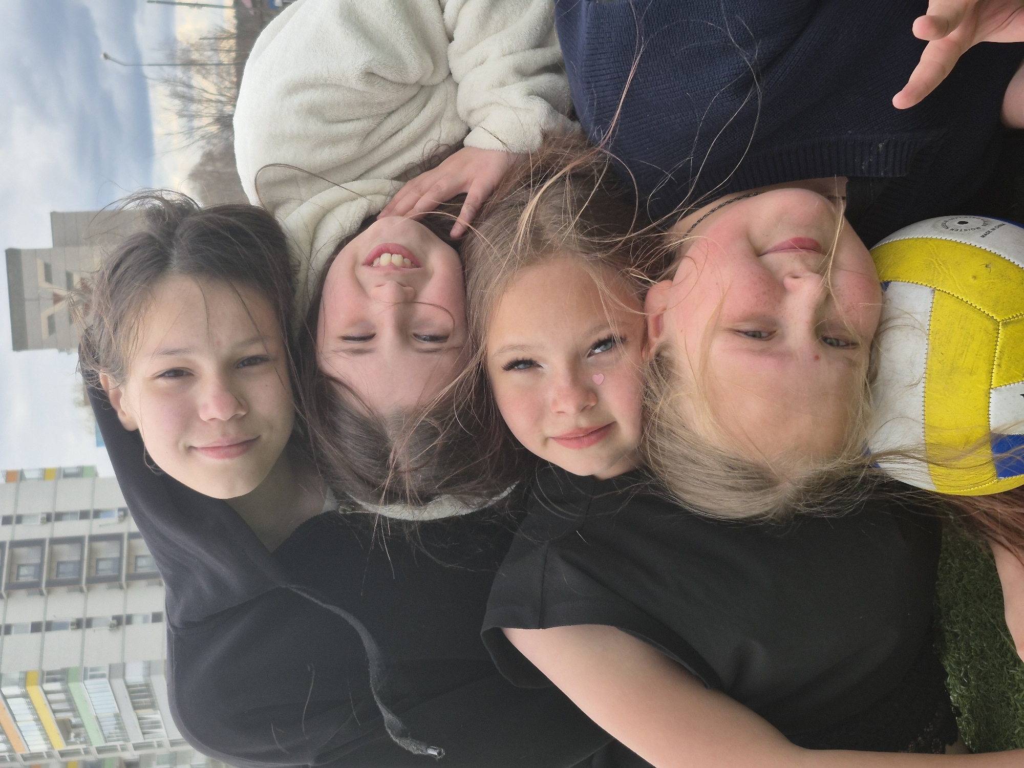 Four young girls are depicted in a top-down outdoor shot, lying on their backs on what appears to be grass, with their heads generally towards the center-right of the frame and bodies extending outwards. The background features tall, multi-story residential or public buildings under an overcast sky.
The girls are arranged in a diagonal line from bottom-left to top-right. The girl at the bottom-left has dark hair and wears a black hooded sweatshirt, smiling subtly. Next to her, a girl with dark hair wears a cream-colored, fuzzy hooded top and smiles broadly, showing her teeth. Adjacent to her, another girl with light brown or blonde hair wears a dark blue knit sweater; she smiles cheerfully and has a small pink heart-shaped sticker near her eye. The girl at the top-right has blonde hair, wears a black t-shirt, and holds a yellow and white volleyball, a portion of which is visible next to her head. The volleyball has text including "VOLLEYBALL", "MOISTURE", and "MADE IN CHINA".
All four girls are smiling, conveying happiness and a sense of companionship. The lighting is soft and even, typical of daytime on a cloudy or overcast day. The buildings in the background are concrete structures with many windows, some featuring distinct colorful panels (yellow, orange, blue, green), suggesting an urban or residential area. The scene depicts a relaxed moment among friends, likely in a park or public space in Chaikovsky, Russia.