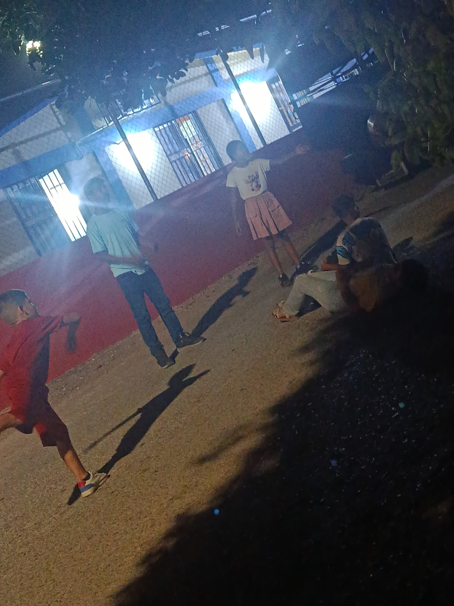 The image shows four children standing on a paved area in Juan Griego, Venezuela. A building with barred windows is in the background, illuminated by bright lights. One child wears a red shirt and red shorts, another a light blue shirt and dark pants, and a third wears a white t-shirt and pink skirt. The fourth child is seated and obscured. Shadows are cast by the children and nearby trees.