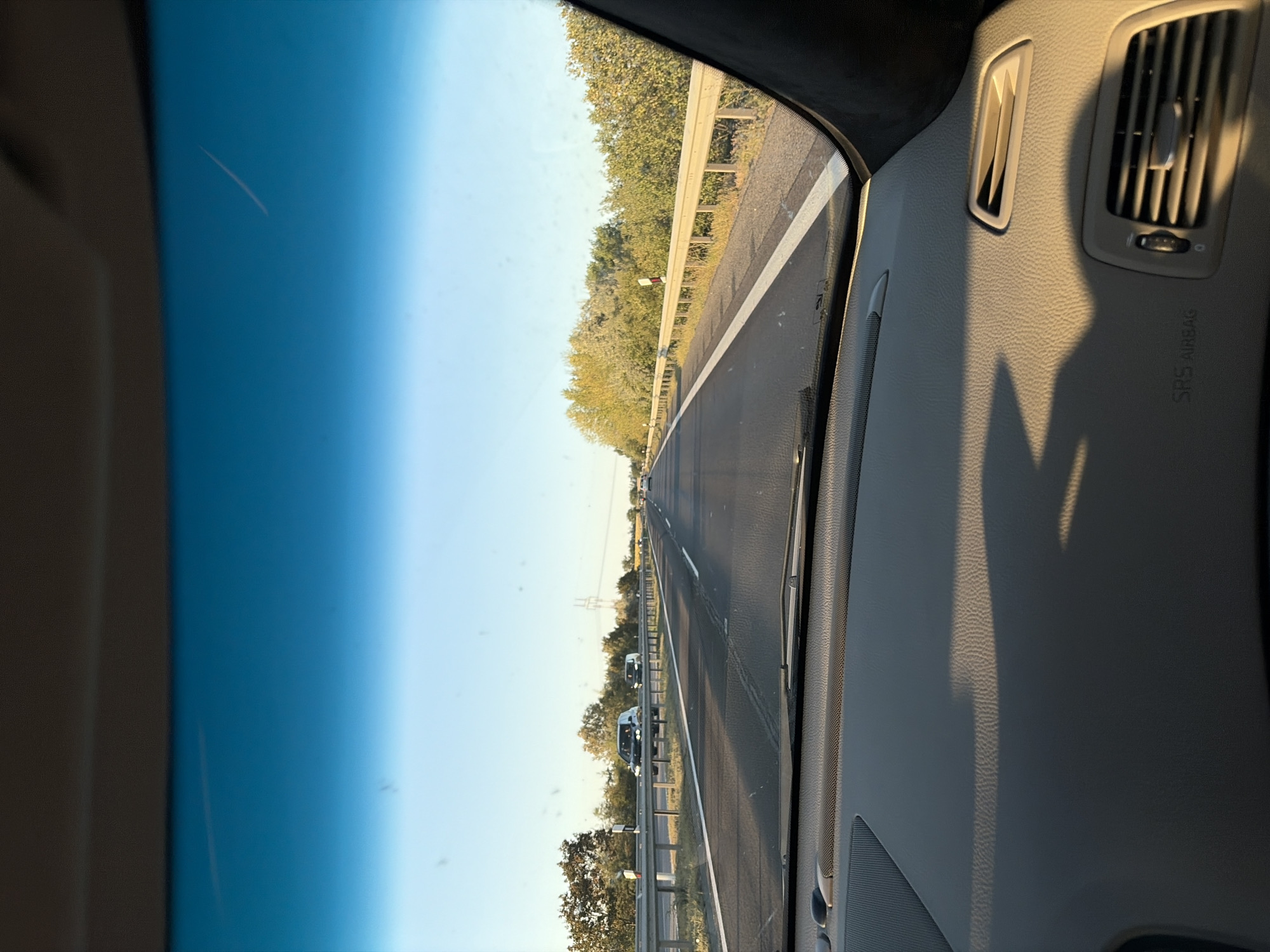 The image is taken from the interior of a vehicle, looking out through the windshield onto a highway. The sun is low in the sky, casting long shadows across the dashboard and the road. The highway appears to be a multi-lane road with a median barrier. There are other vehicles on the road, including cars and vans. Trees and vegetation line the sides of the highway. In the distance, power lines and poles are visible. The sky is a clear blue with a faint contrail from an airplane. The dashboard of the car is visible in the foreground, with an air vent and "SRS AIRBAG" lettering. The scene depicts a drive on a highway during a sunset, likely in a rural or suburban area, given the presence of vegetation and the open expanse of the road. The specific location in Pata, Slovakia cannot be determined from the visual information alone, as there are no identifying landmarks or signs visible in the image.