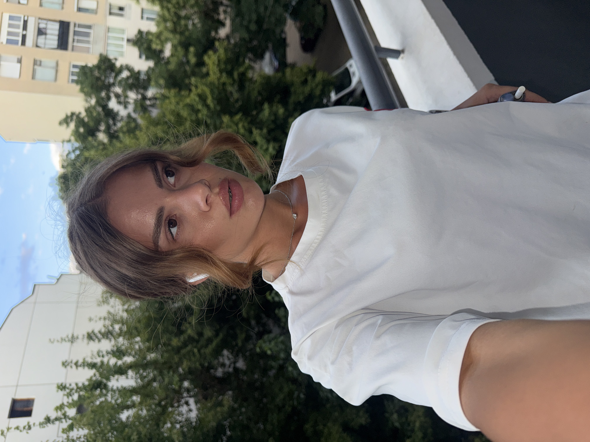 A young woman stands on a balcony, holding her phone up to take a selfie. She is wearing a white t-shirt and has on white earbuds. Her hair is pulled back and styled in curls. The background shows trees and buildings, suggesting an urban environment. The woman is wearing a ring on her finger. The scene appears to be in Plovdiv, Bulgaria, based on the architecture of the buildings in the background.