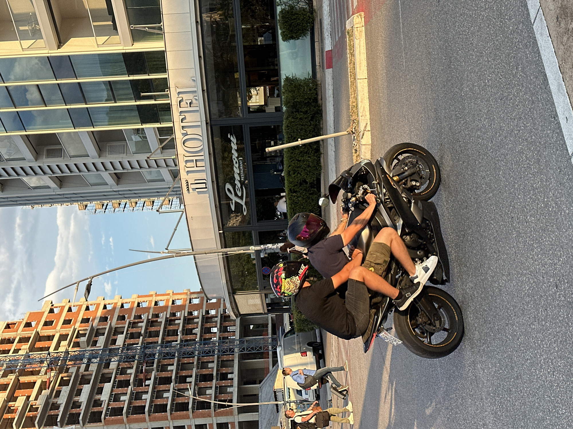Two individuals wearing helmets are riding a black motorcycle on an asphalt road in Budva, Montenegro. The person in front is driving, while the person behind is a passenger. Both are wearing casual clothing. The motorcycle is equipped with a license plate. In the background, there is a building under construction, characterized by exposed brick and a crane. Adjacent to it, a modern hotel building with large glass windows and balconies is visible, with a sign that reads "HOTEL" and "Lemoni Restaurant". The scene is illuminated by natural sunlight, casting shadows. The road also features pedestrian crossing markings.
