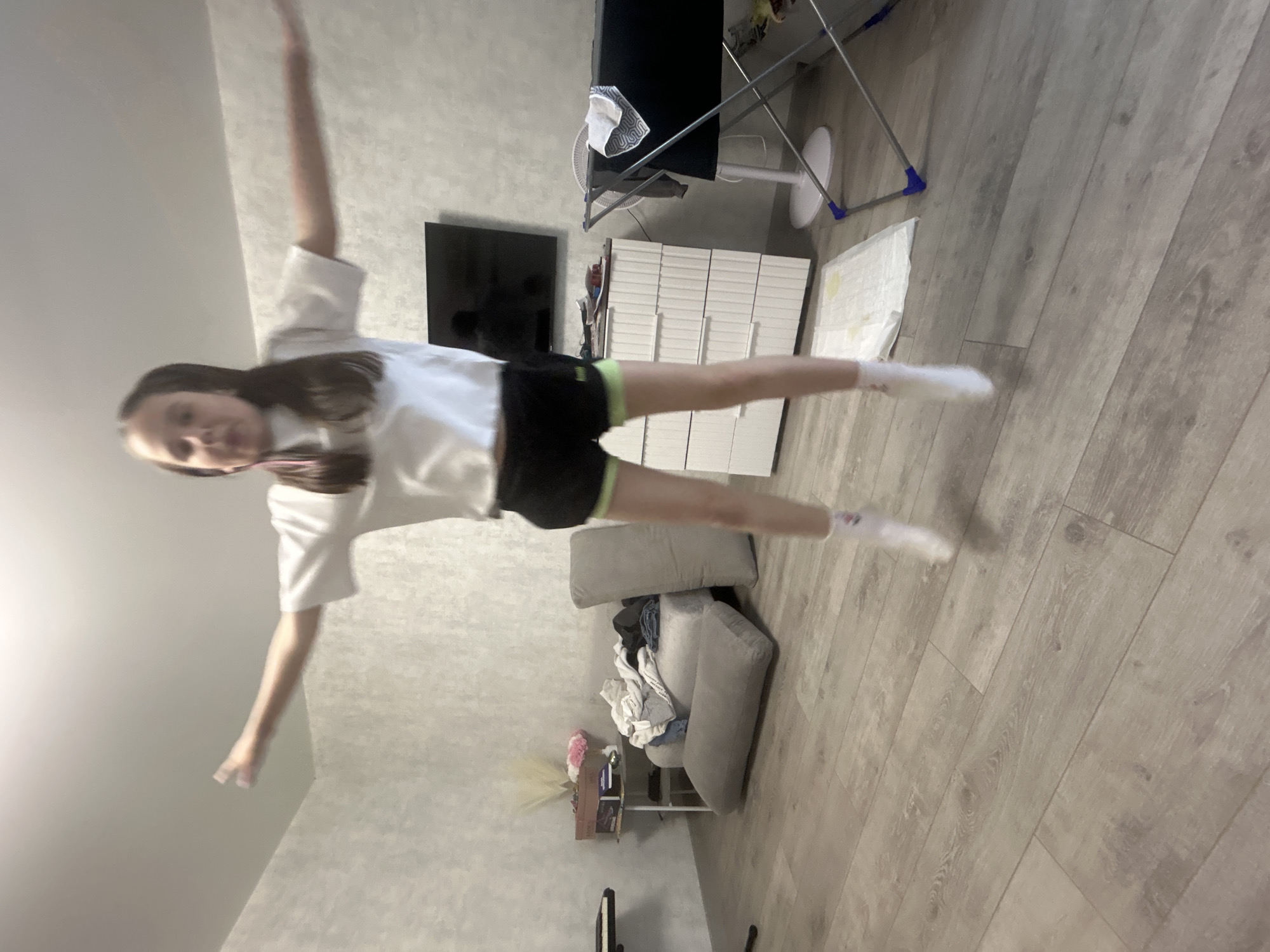A young girl with brown hair, wearing a white t-shirt and black shorts with neon green trim, jumps in the air with her arms outstretched. She is wearing white socks. The scene appears to be an indoor residential setting, possibly a living room or bedroom. The room has light-colored walls and a wood-plank floor. In the background, there is a sofa with a pile of clothes on it, a chest of drawers, a television, and a drying rack with a fan. The overall impression is of a casual, everyday moment captured in a domestic environment.