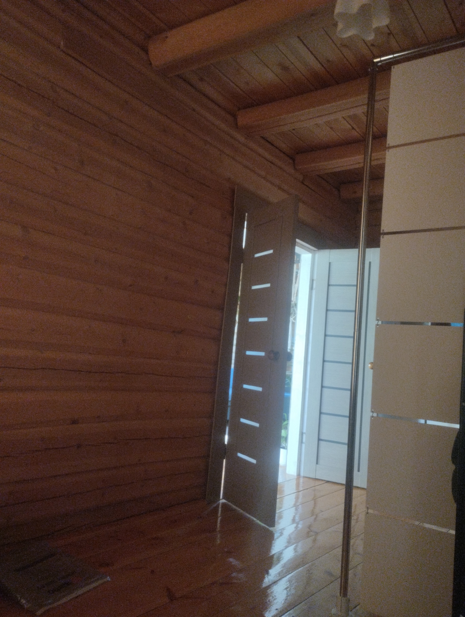 The image depicts the interior of a room with wooden walls and ceiling. A highly polished wooden floor reflects the light. In the foreground, a door is partially open, revealing another door beyond it. A metal pole runs vertically through the center of the image, separating the two doors. The room appears to be under construction or recently renovated, with a somewhat unfinished look. The image does not provide any specific visual cues to determine its location within Андреевский, Russia, or any context regarding people, actions, or interactions.