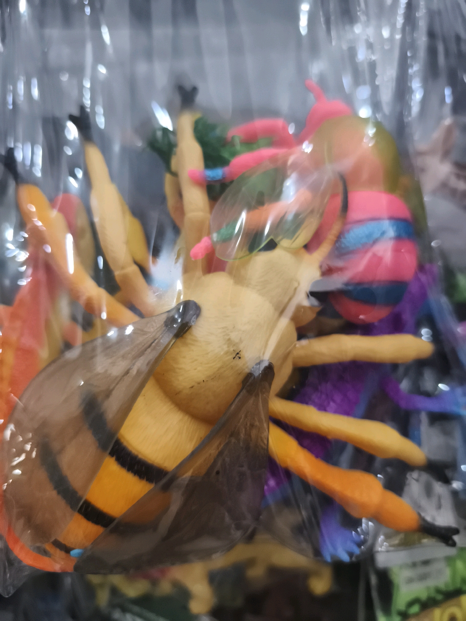 The image shows a close-up of several colorful toy insects, likely made of plastic, contained within clear plastic packaging. The toys are brightly colored with prominent yellow and orange stripes on the largest insect, which appears to be a bee or wasp. Other toy insects in various colors like pink, purple, green, and blue are also visible. The lighting suggests an indoor environment, possibly a store or market stall, with bright reflections on the plastic packaging. The background is blurred and indistinct, but hints of other merchandise are visible. The specific location in Palembang, Indonesia, is not identifiable from the image itself.