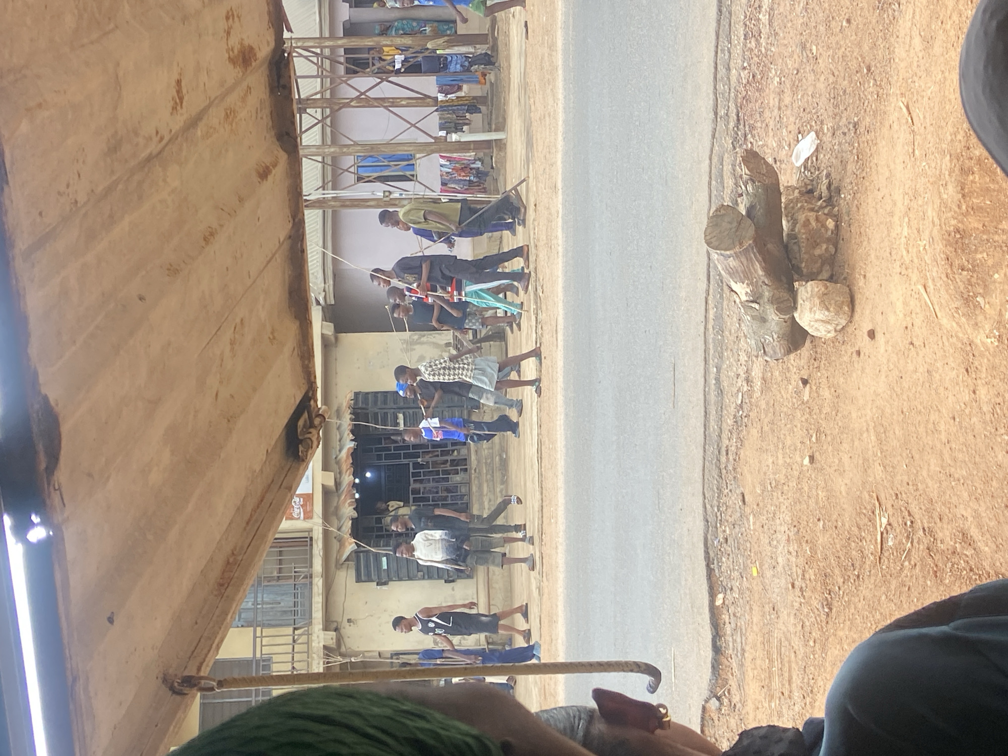 A ground-level, slightly tilted view from within a vehicle or shelter in Okengwe - Obehira, Nigeria, shows a group of people walking along a road. The group comprises primarily young men and boys. They are moving from right to left in the frame along the left side of the paved road. Some are carrying long, thin objects, possibly sticks. The buildings lining the road on the left side have a worn, urban look. Clothes are hung out to dry on balconies or structures attached to the buildings. On the right side of the road, the ground is unpaved with a dirt surface. A pile of logs and rocks sits on the roadside. The view is partially obstructed by the roof of the structure, which has a metallic or corrugated appearance. The lower part of the frame includes a glimpse of an individual wearing green clothing.