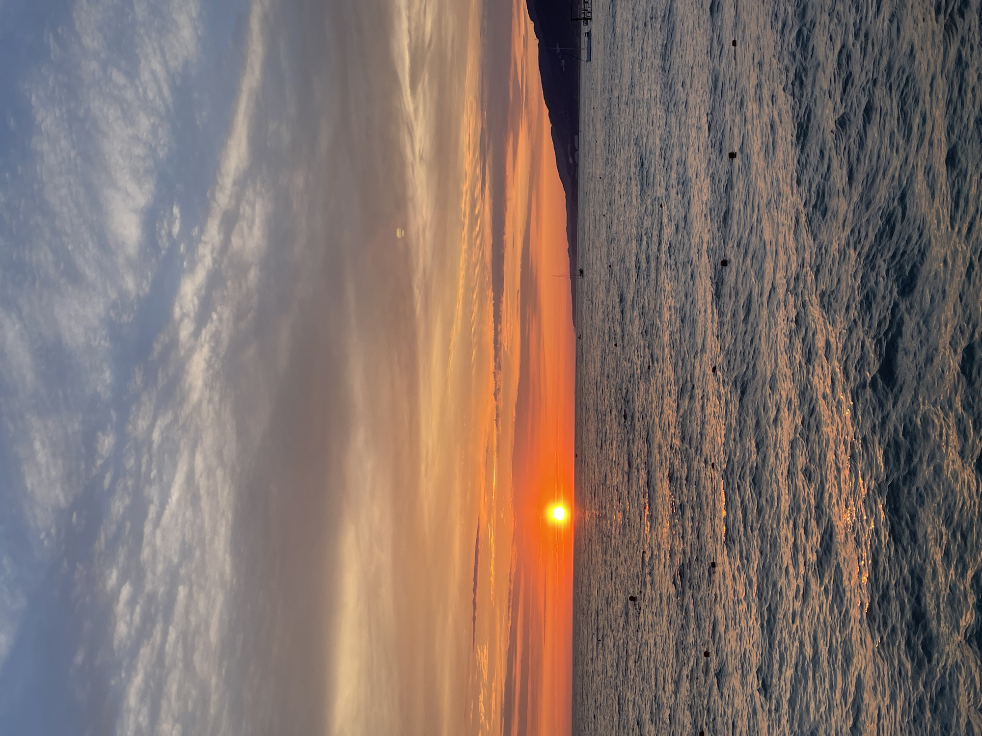An outdoor, full shot captures a vibrant sunset over the ocean. The sun, a bright orange orb, is positioned in the center-left of the image, casting a warm glow across the sky and water. The sky transitions from a soft yellow and orange near the horizon to shades of pink and blue at the top, with scattered, wispy clouds. The ocean in the foreground is a deep blue, with gentle waves reflecting the sunset's colors. Dark buoys are visible scattered across the water. In the distance, a dark, mountainous coastline is silhouetted against the colorful sky. A few boats are present on the water, including a sailboat near the shore. A pier with some structures is visible on the right side of the distant coastline.