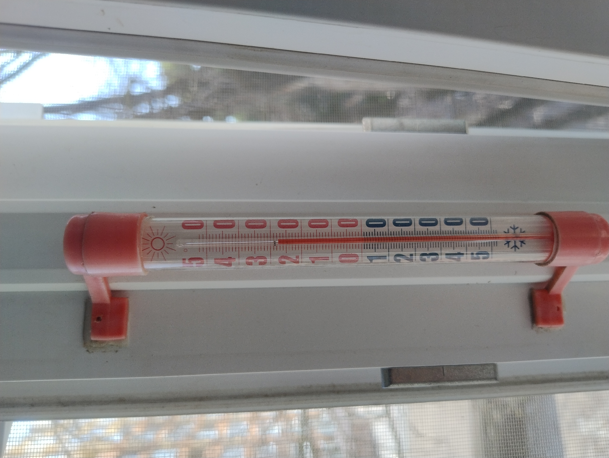 An outdoor thermometer is attached to a white window frame. The thermometer is red and white and displays temperature in Celsius. The liquid level in the thermometer is at 1 degree Celsius. The surrounding environment appears to be outdoors, with a blurry view of trees and branches visible through the window.