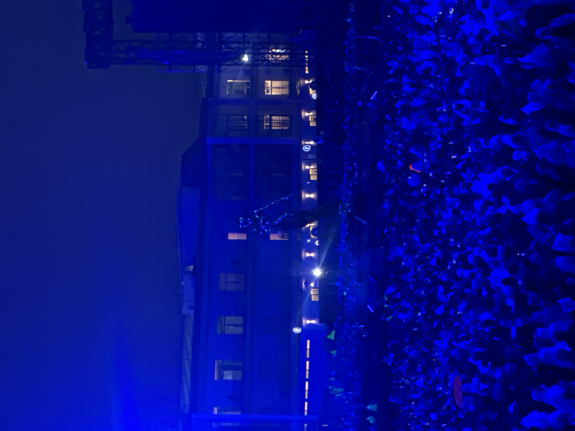 The image captures a night scene in Mexico City, Mexico, featuring a large crowd illuminated by blue light, possibly attending an event or gathering. To the left, a tall building is visible with lit windows and architectural features. The crowd is densely packed, with individuals distinguishable only by their silhouettes in the intense blue lighting. The overall environment suggests an urban setting, likely a public space or event venue. The atmosphere is vibrant, with a focus on the crowd's presence and the building's backdrop.