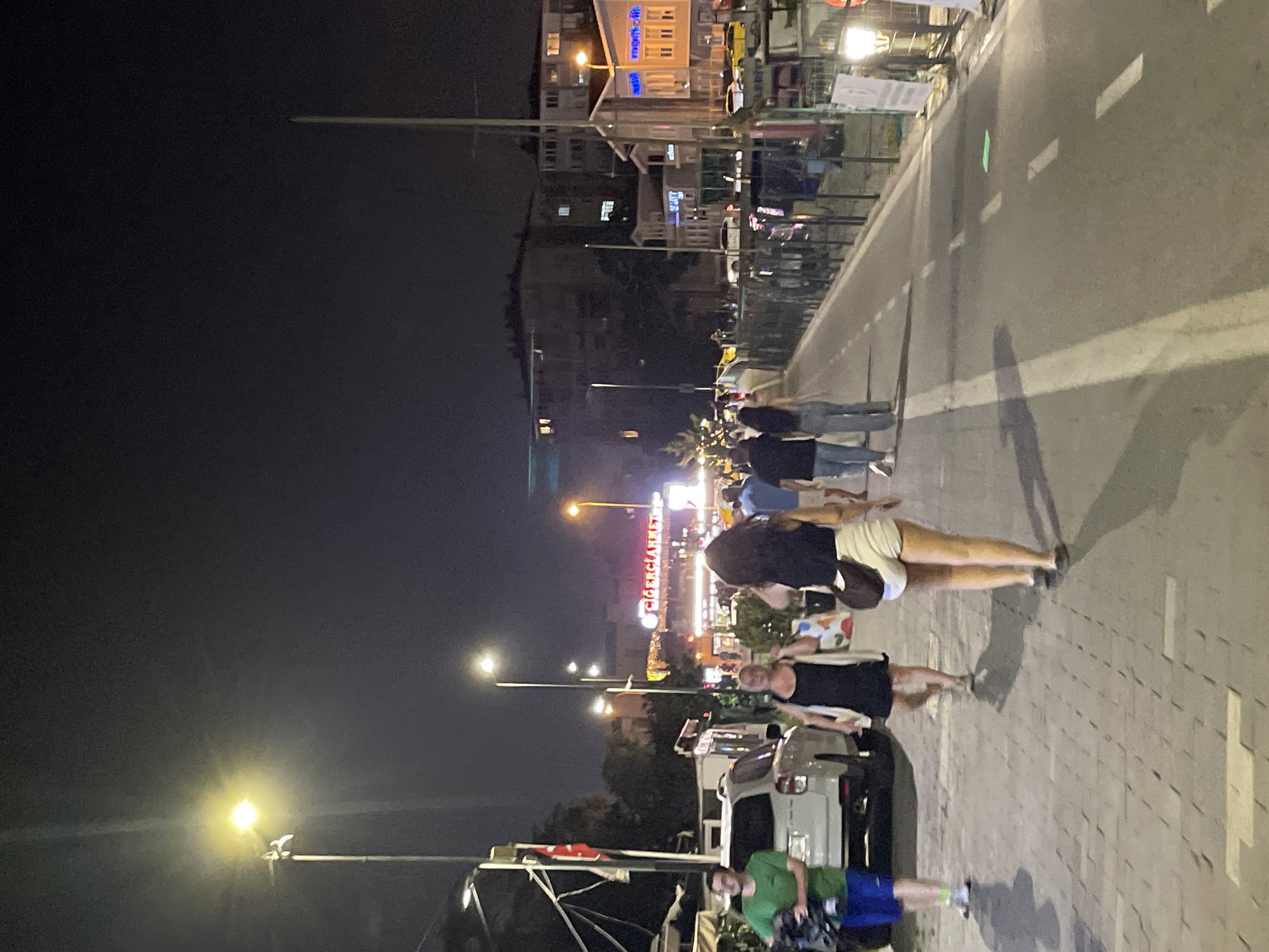 An exterior, nighttime shot captures a bustling pedestrian walkway in Kadıköy, Turkey. The scene is illuminated by streetlights and the lights from various shops and businesses lining the walkway. Several people are walking in both directions, some appearing to be tourists or locals enjoying a night out. On the left side of the walkway, a white SUV is parked, with a person in a green t-shirt and blue shorts standing next to it. Further down the walkway, a woman in a black dress and a cardigan is walking away from the camera, followed by a woman in a black tank top and white shorts. In the background, buildings with illuminated windows suggest a vibrant urban environment. The walkway is paved with cobblestones and asphalt, with distinct white lines marking a lane. The overall atmosphere is lively and social, indicative of a popular area in Kadıköy.