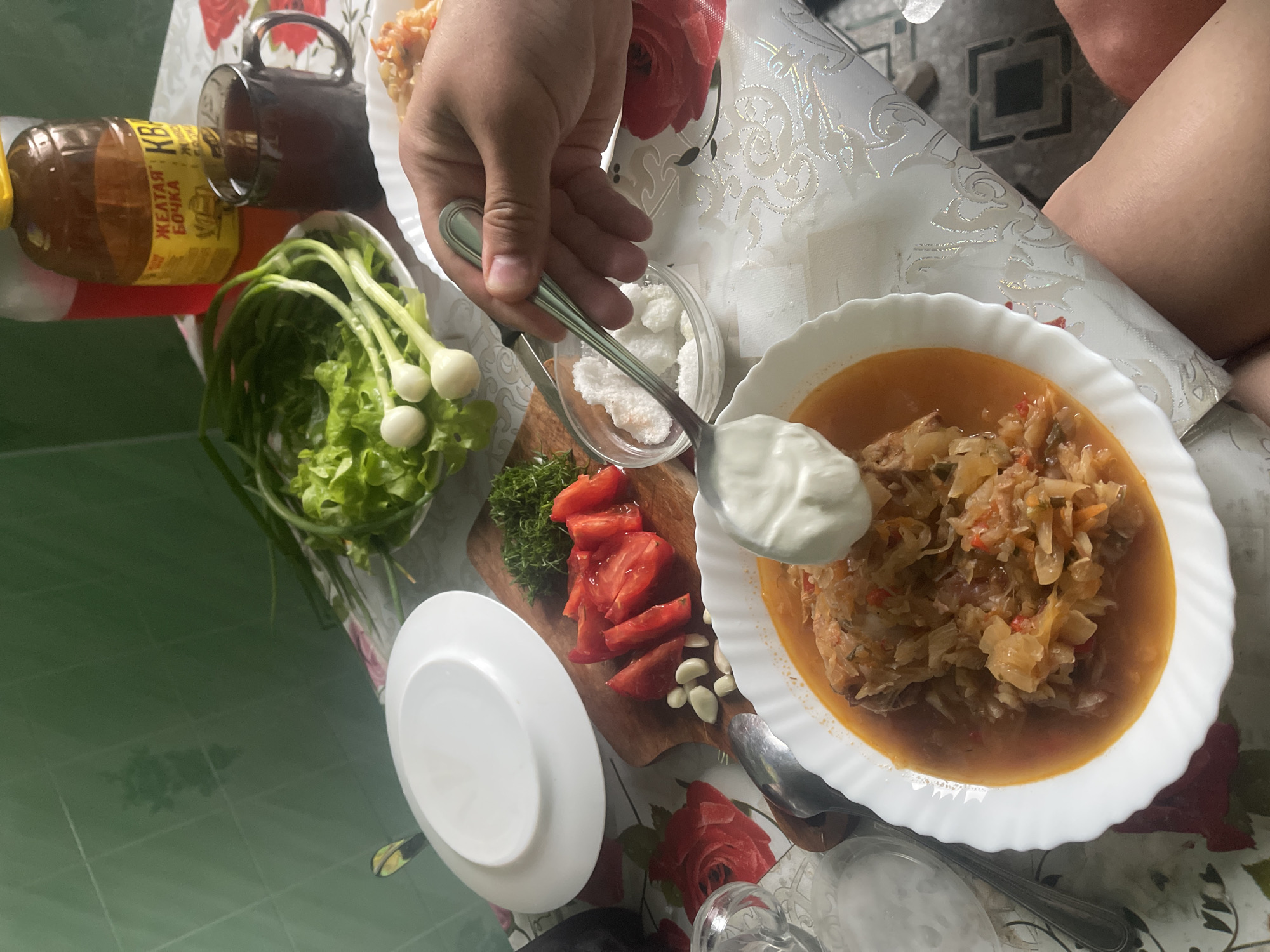 An overhead shot captures a table laden with food, suggesting a meal in progress at an indoor location, likely a home, in the city of Krasnoe, Russia. The table is covered with a white tablecloth patterned with red roses and gold accents. In the foreground, a white bowl holds a stew of chopped cabbage and meat, topped with a dollop of sour cream. A spoon, held by a person's hand, is poised to add more sour cream. Beside the bowl, a small glass dish contains what appears to be cubed white cheese or sugar. To the left, a cutting board displays sliced tomatoes, garlic cloves, and a sprig of dill. Further back, a plate of fresh green lettuce and scallions sits near a dark mug of tea and two bottles, one with red liquid and another amber bottle labeled in Cyrillic. The background features a green tiled wall.