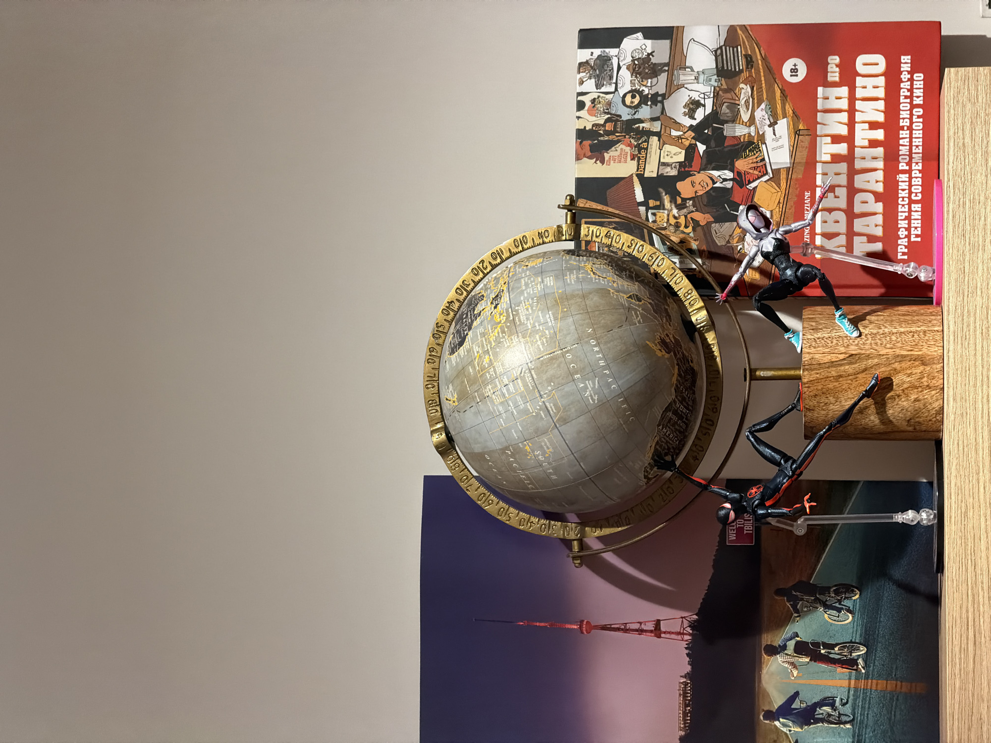 The image shows a globe with a golden meridian ring, displaying "North Pacific" on its surface. Leaning against it is a book with a cover featuring the Tbilisi TV Broadcasting Tower and cyclists. There are two action figures of Spider-Man characters posed in front of the globe and books. One is Spider-Gwen, in a dynamic pose. The other is Miles Morales, also posed mid-air. The globe, books, and action figures are on a wooden surface. One of the books with a red cover displays "Квентин Тарантино" ("Quentin Tarantino") in Russian. The other book is a comic book.