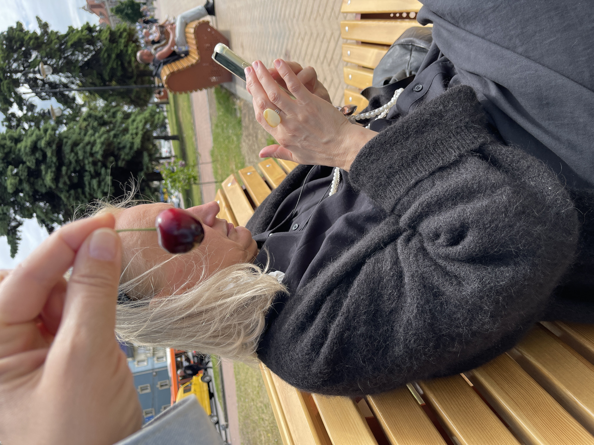 A woman with white hair is sitting on a wooden bench outdoors, using her mobile phone. She is wearing a black cardigan, a black button-down shirt, and a pearl necklace, and she has a ring with a large, light-colored stone on her right hand. Another hand is holding a dark red cherry on a stem in front of the woman's face.
The background includes a paved walkway, green trees, and a building with a red roof and white trim. There is a sculpture of a man playing an instrument seated on a bench. Based on the elements present, the location is likely within the city of Kaliningrad, Russia.