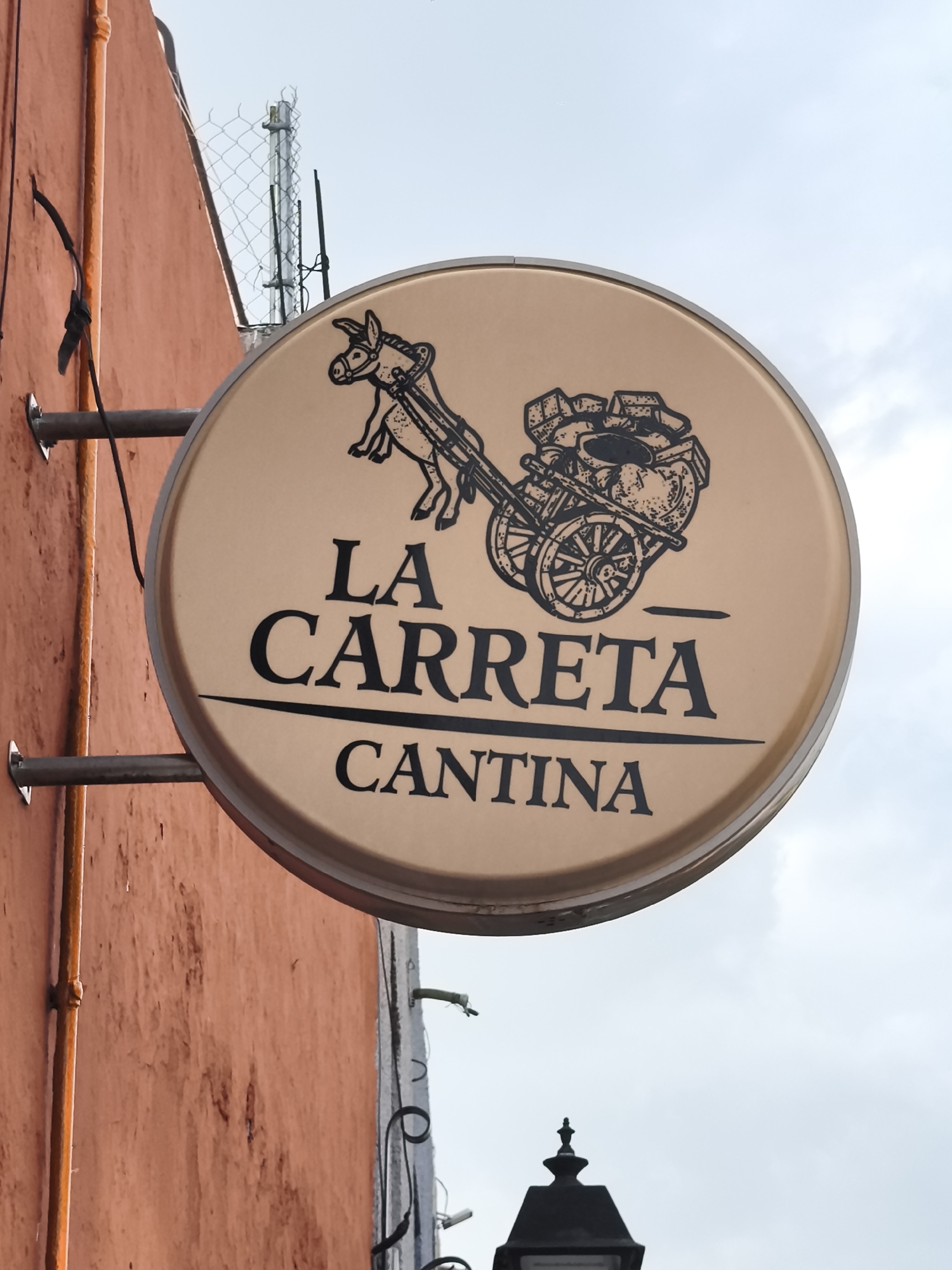 A round, illuminated sign for "La Carreta Cantina" hangs on the side of a red-painted building. The sign is light brown with black lettering and a cartoon donkey pulling a cart. The sign is attached to the building via two horizontal metal bars. The wall is textured and shows signs of wear. A vertical orange pipe is affixed to the wall near the sign. In the background, there is a wire fence and a partly visible street lamp. The sky is overcast. This is likely located in the city of Querétaro, Mexico as indicated by the name of the cantina.