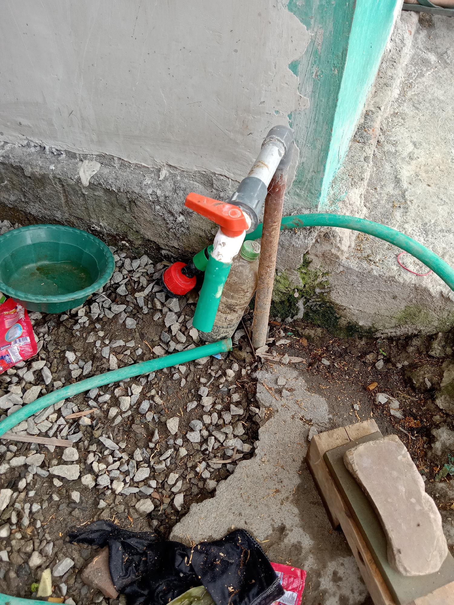 The image depicts an outdoor scene at ground level, likely in a residential area in Pacing, Indonesia. In the center, a green garden hose is connected to a faucet with an orange handle and a white and grey pipe assembly. Next to the faucet, a red nozzle is attached to the hose. To the left of the faucet, a green basin is partially filled with water and rests on a bed of small grey stones and dirt. A red packet, possibly detergent or soap, lies near the basin. Behind these items, a textured concrete wall forms the backdrop, with patches of green paint visible on the right side. The ground is covered with gravel, dirt, and some fallen leaves. In the lower right corner, several wooden planks are stacked, with a flat, light-colored stone placed on top. The overall impression is of a functional, utilitarian outdoor space, possibly a backyard or a side yard.