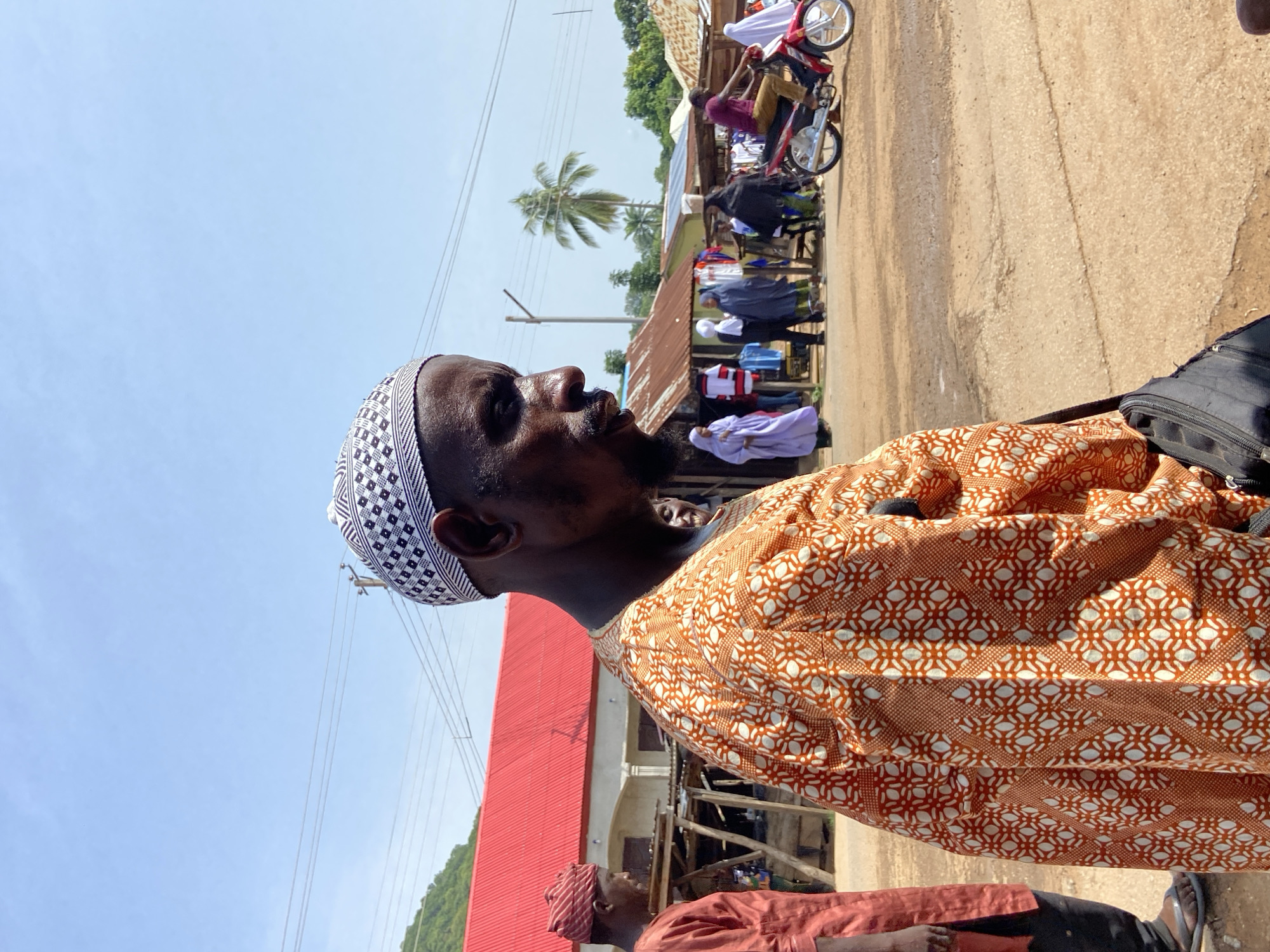 A medium shot shows a man in an orange patterned shirt and a white and black kufi cap standing outside in what appears to be a street in Mararaba, Nigeria. The man has dark skin, a beard, and is looking to the right. In the background, there are other people, some wearing white robes, and a person on a red motorbike. There is a red-roofed building and a palm tree visible in the background. The ground is dirt or unpaved. The sky is light blue and clear.