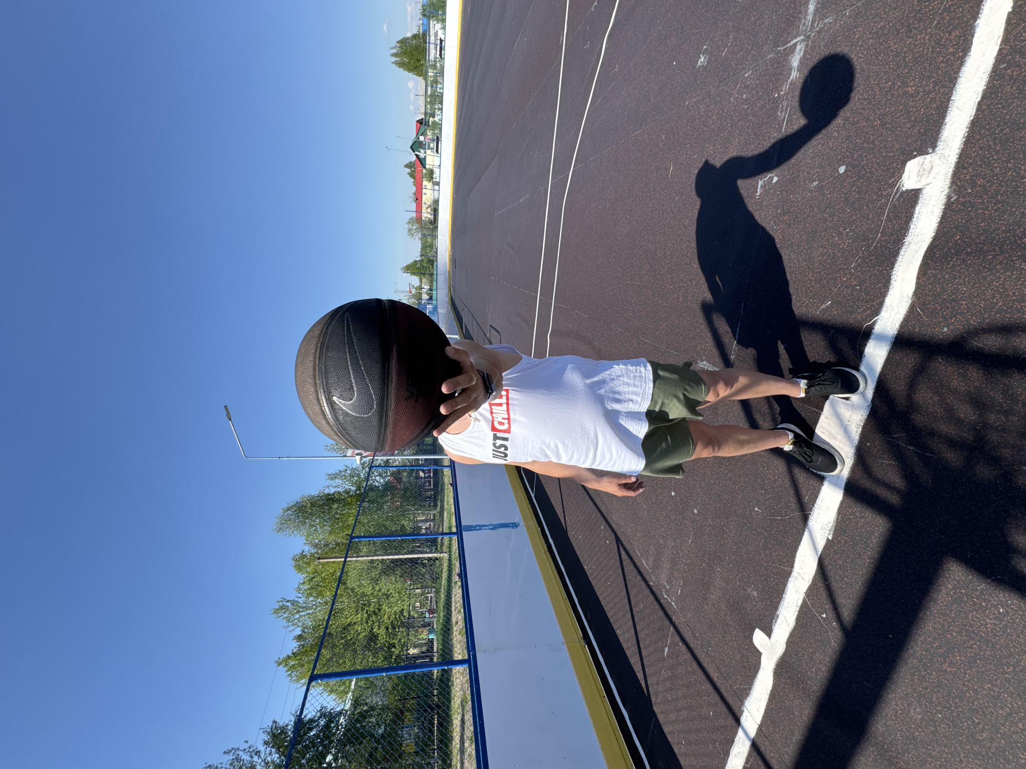 A vertical, eye-level photograph shows a person standing on an outdoor sports court in Vysokiy, Russia, on a bright, sunny day. The individual is wearing a white sleeveless top with the red text "JUST CHILL" printed on the chest, and olive green shorts. They are holding a black basketball, emblazoned with a white Nike swoosh, directly in front of their face, completely obscuring it. Dark sneakers with white soles are visible on their feet.
The court surface is dark, likely asphalt or a rubberized material, clearly marked with crisp white lines, indicating it's a basketball or multi-sport facility. Strong sunlight casts a distinct shadow of the person and the basketball on the court to the right. The sky overhead is a brilliant, cloudless blue, indicating a clear weather condition. In the background, a blue chain-link fence runs along the side of the court, behind which a lush line of green trees and distant residential buildings can be seen. A tall street light pole extends into the blue sky on the left. The scene conveys a relaxed outdoor moment on a well-maintained court.