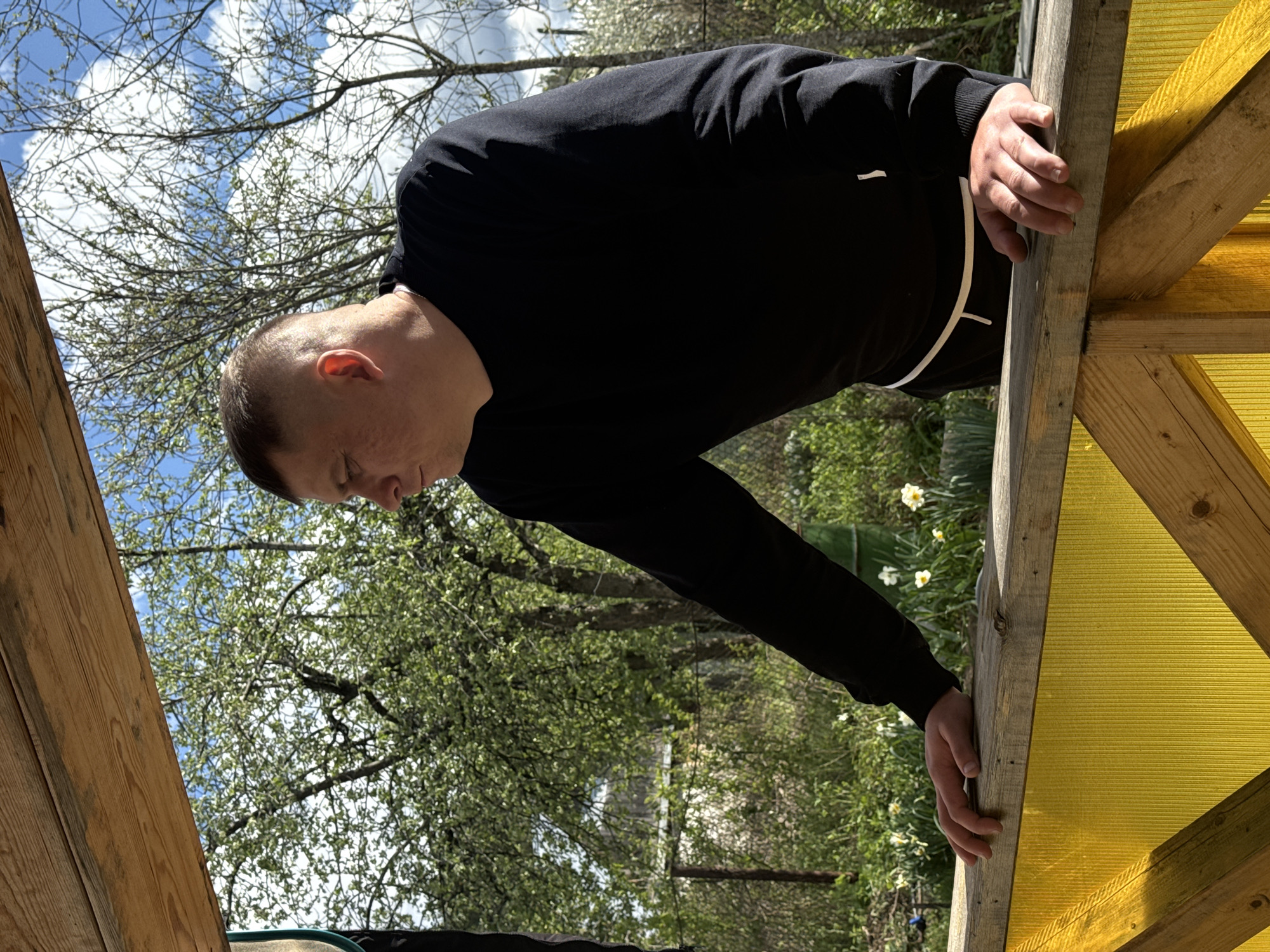 A man wearing a black sweater and white trim around the waist is bent over and holding onto the wooden supports of a structure with yellow transparent panels. He has short hair and appears to be looking down. Behind him are trees and a blue sky with white clouds. The setting includes blooming white flowers and a glimpse of what seems to be a building in the background. It is daytime. The image was taken outdoors in the city of Chaikovsky, Russia.