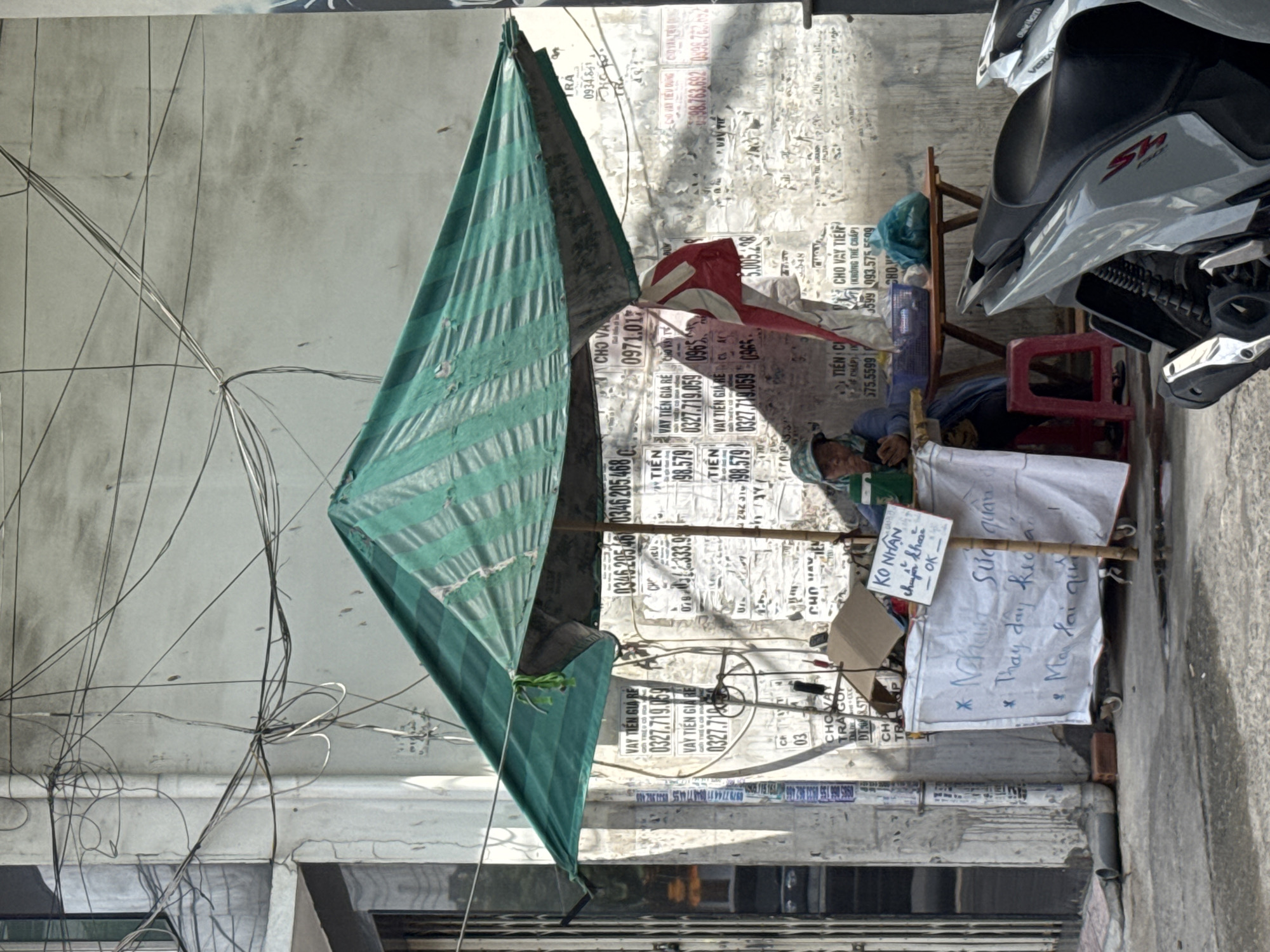 The image shows a street scene in Nha Trang Ward, Vietnam. A person is seated at a makeshift stall under a green tarp, appearing to be a vendor. The stall consists of a white cloth-covered table on wheels, with various items on it, including a cardboard box and some papers. A small sign with Vietnamese writing is attached to a bamboo pole. Behind the stall, a wall is covered with numerous posters and advertisements. A wooden table and stool are also visible, along with some bags. A motorcycle is parked in the foreground. The environment suggests an outdoor commercial area, likely a street market or roadside stall. The wall and the abundance of posters indicate a densely populated urban setting where advertising is common. The overall impression is one of everyday commerce in a Vietnamese city.