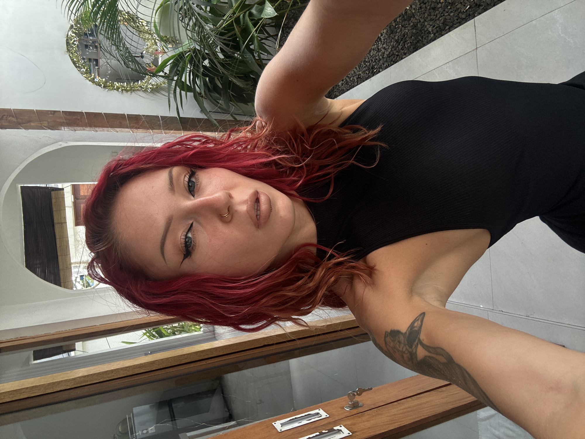 A medium shot of a Caucasian woman with red hair, wearing a black sleeveless top, and facing the camera. She has a nose ring and her arm, which has a tattoo, is visible. The background includes a building with windows, a wooden frame, and a small key near the bottom. A round mirror with a decorative border and plants are visible in the upper left corner. The location is identified as North Kuta, Indonesia.