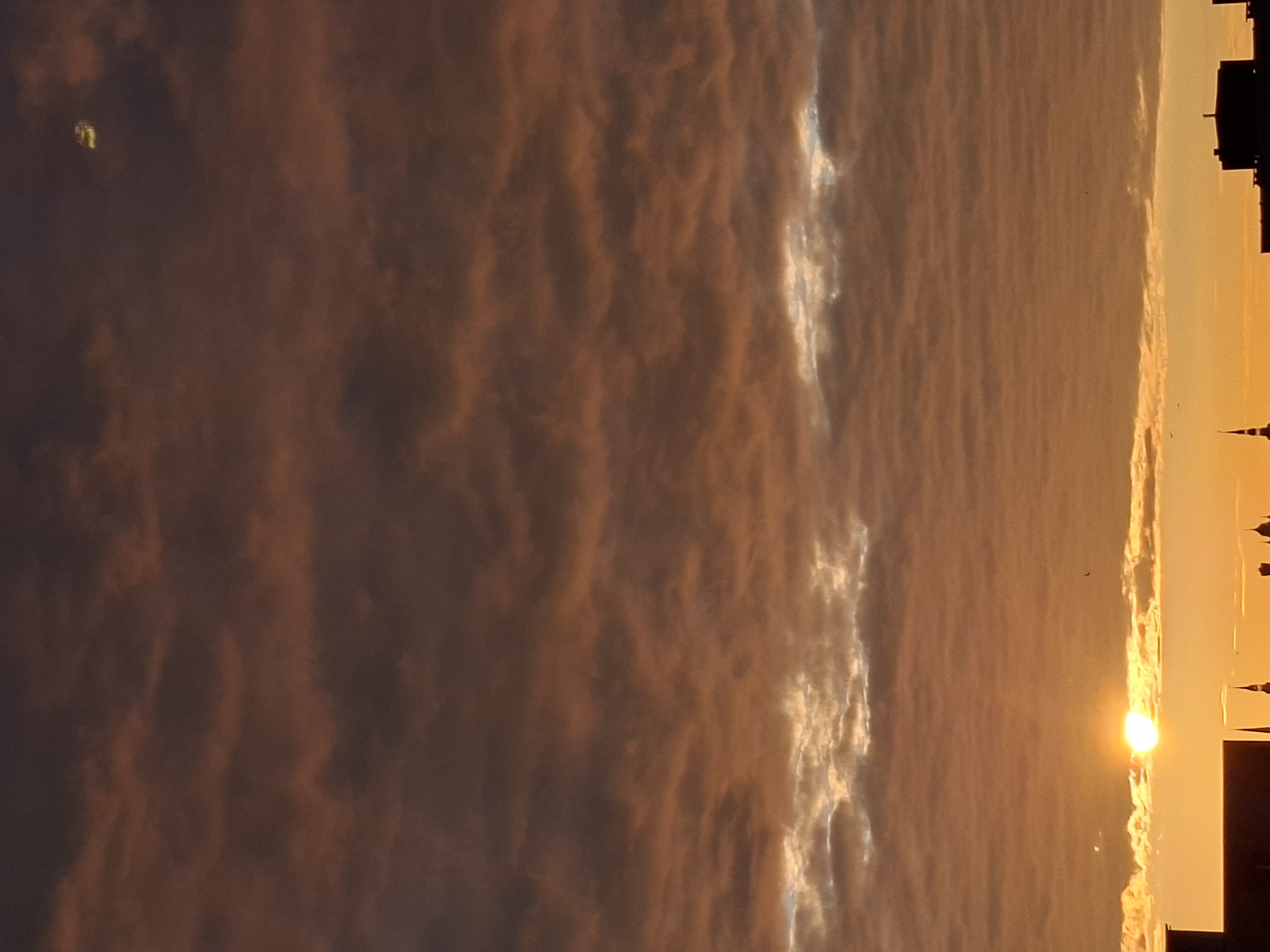 The image displays a vibrant sunset or sunrise over Tallinn, Estonia, dominated by a sky filled with dramatic cloud formations. The photograph is oriented vertically, but if viewed horizontally, the sun would be on the right side, illuminating the scene from a low angle.
The sky is largely covered by layered, textured clouds in rich tones of orange, reddish-brown, and dark gray. Patches of these clouds are brightly illuminated by the sun, appearing as brilliant streaks and glowing sections of silver-gold. These contrasting lights and shadows emphasize the dynamic structure of the cloud cover.
The sun is visible as an intense, bright orb, partially obscured by the atmosphere or clouds, casting a strong golden light across the horizon. Beneath this glowing horizon, the dark silhouettes of a city skyline are visible, featuring indistinct buildings, some with discernible spires or towers.
In the upper left portion of the sky (when viewed horizontally), a small, blurry, yellowish-green object or light anomaly is present, its exact nature unclear. The overall atmosphere is one of natural grandeur and tranquility, capturing the beauty of a twilight sky over an urban landscape. There is no visible text in the image.