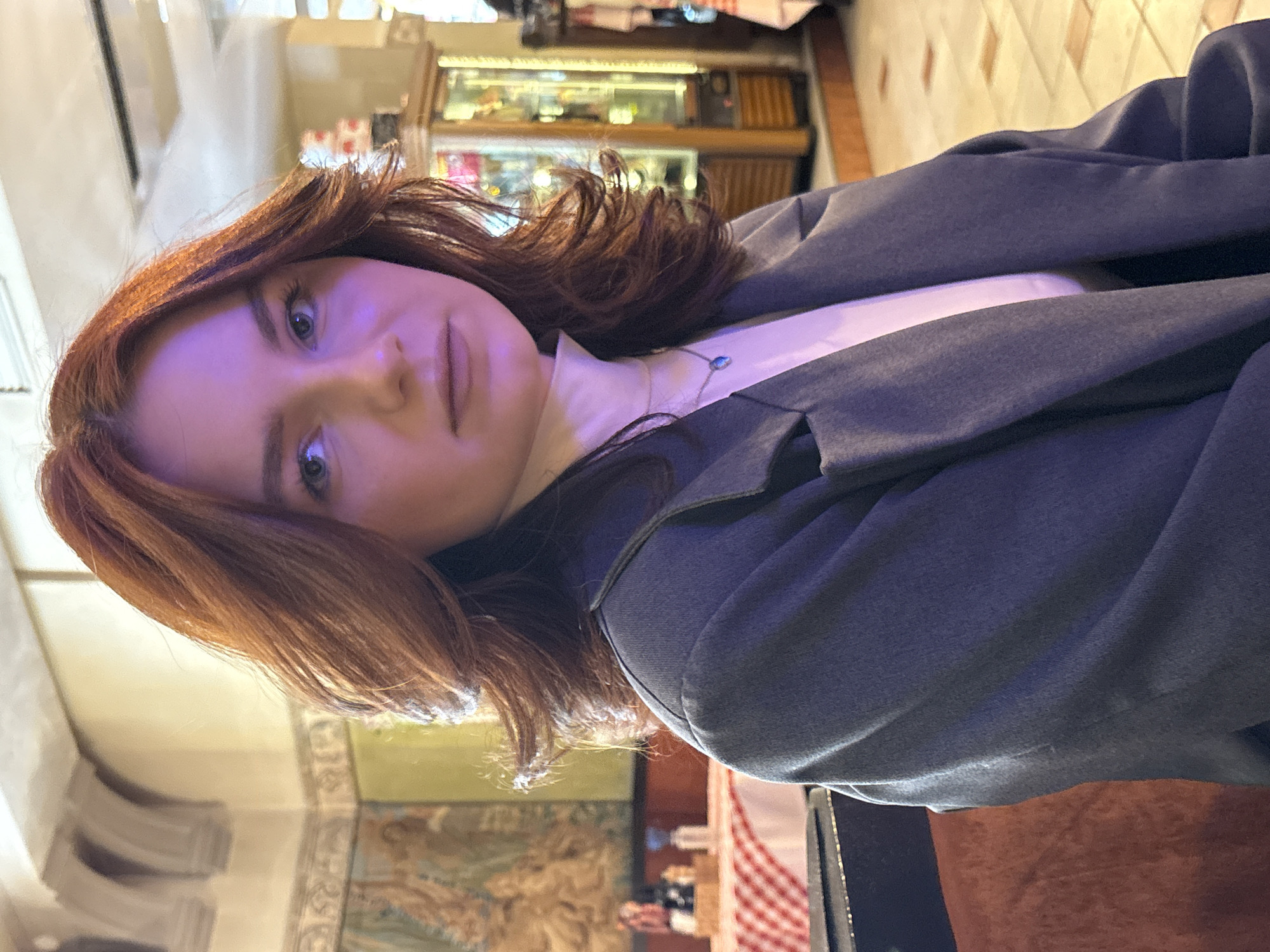 The image shows a woman with auburn hair wearing a gray jacket and a white top. She is positioned indoors, with a blurred background featuring restaurant decor, including tables with red and white checkered tablecloths and a wall mural. In the upper background, there is a glass display case. The setting appears to be a restaurant, possibly in Saint Petersburg, Russia, given the style of interior design.