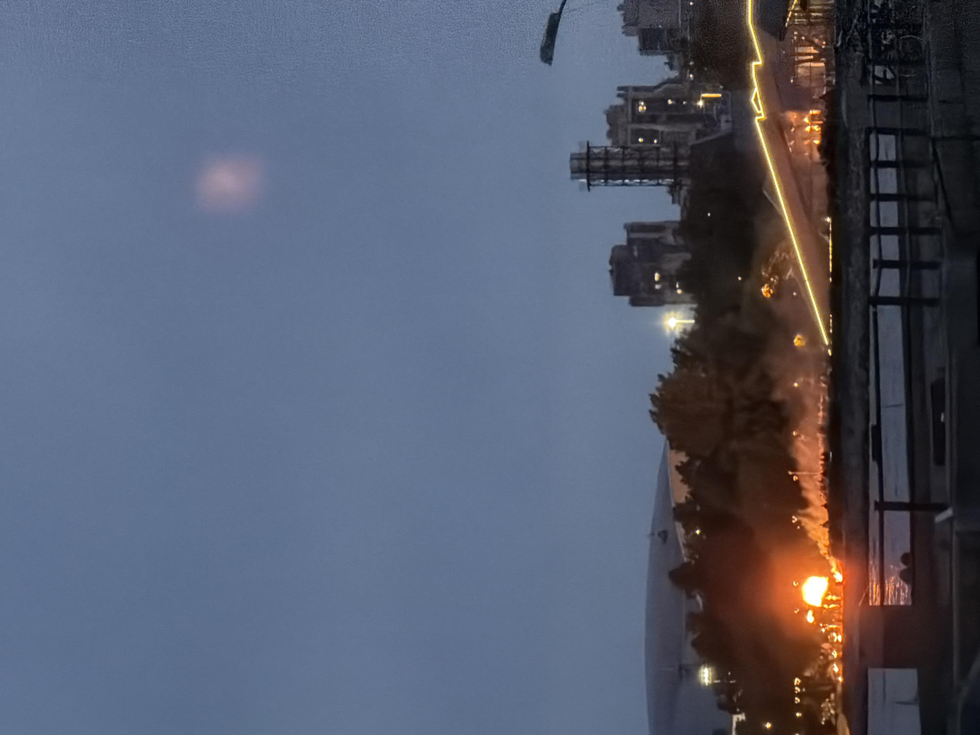 The image depicts an industrial area at dusk, with a prominent fire in the foreground. The fire emits a bright orange glow and a large plume of smoke that rises into the dim, blue sky. Silhouettes of people are visible near the fire, suggesting a gathering or event. In the background, several industrial buildings and structures, including towers, are illuminated by lights. A faint, pinkish glow is visible in the sky above the industrial complex. A street light on the right side of the image casts a dim light. The scene is framed by a dark foreground element, possibly a railing or bridge, with a body of water below reflecting the ambient light. The overall atmosphere is one of industrial activity and a contained fire, set against a backdrop of an encroaching night.