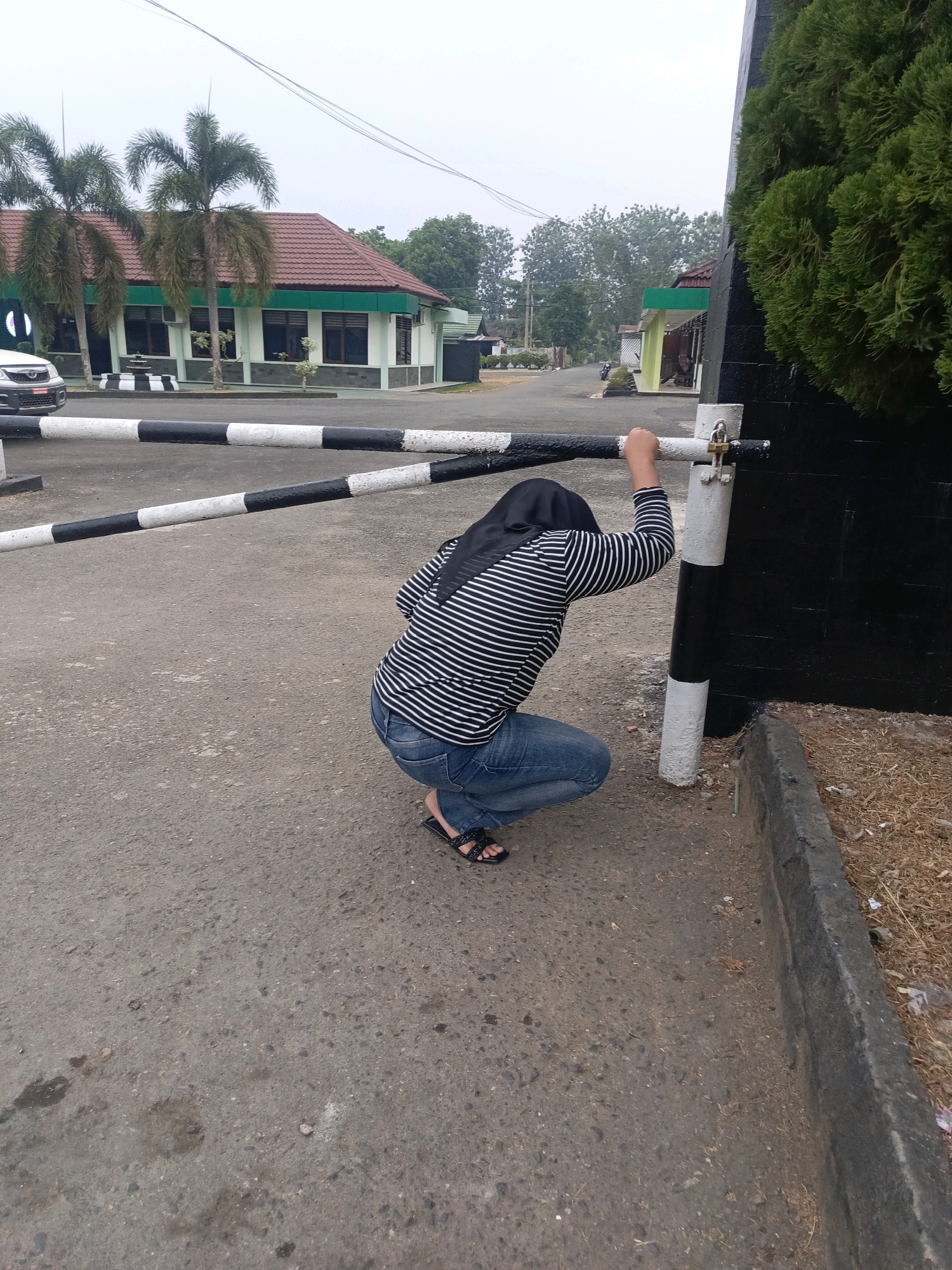 A person wearing a black and white striped long-sleeved shirt, blue jeans, and black sandals is squatting next to a black and white striped barrier pole. The person is wearing a black headscarf and has their right hand on the barrier pole, which is locked with a padlock. The barrier is part of a larger gate that appears to be at an entrance to a building complex. In the background, there is a light-colored building with a red roof, palm trees, and other vegetation. The ground is paved with asphalt. The image appears to have been taken outdoors in a relatively bright but overcast environment. The context of Lubuklinggau, Indonesia suggests a tropical or subtropical climate.