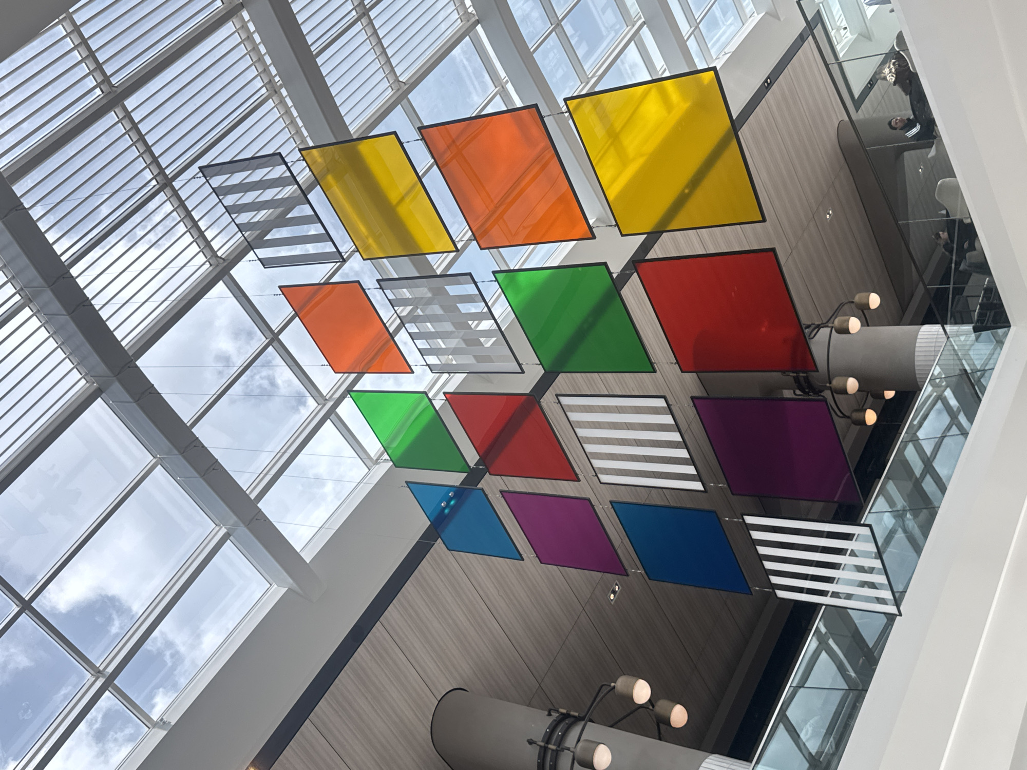 The image shows a view looking up at an array of colorful, diamond-shaped panels suspended from the ceiling of an indoor space. The panels are arranged in rows and feature various colors including yellow, orange, red, green, blue, and purple, interspersed with panels featuring horizontal white stripes. The ceiling above is a grid of metal beams and glass panels, allowing natural light to filter through. Part of a second-level balcony or walkway can be seen to the right of the frame, along with what appears to be a cylindrical support structure with light fixtures attached. The overall environment suggests a modern, open architectural design. I do not have enough information to determine where it is located.