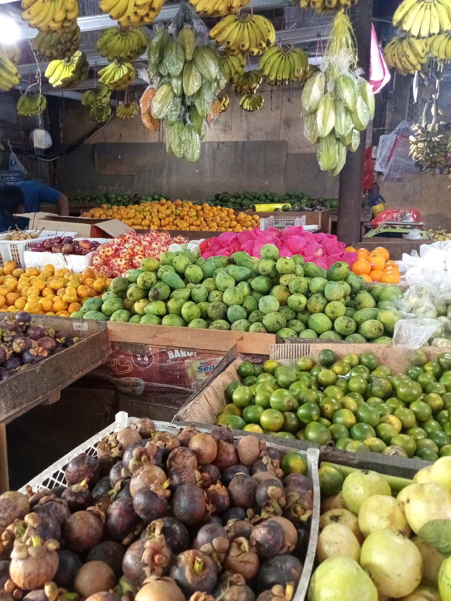 The image depicts an outdoor fruit stall in Setiamekar, Indonesia. Bunches of yellow bananas and green starfruit hang from the ceiling. In the foreground, various fruits are displayed in crates and on tables. To the left, a crate is filled with mangosteens. Adjacent to this, a large pile of green mangoes is visible. Further back, there are piles of oranges and smaller green fruits. A person in a blue shirt is partially visible in the background, appearing to be working at the stall. The stall is set up against a simple wall, and other fruits are displayed in the background as well, suggesting a bustling market environment.