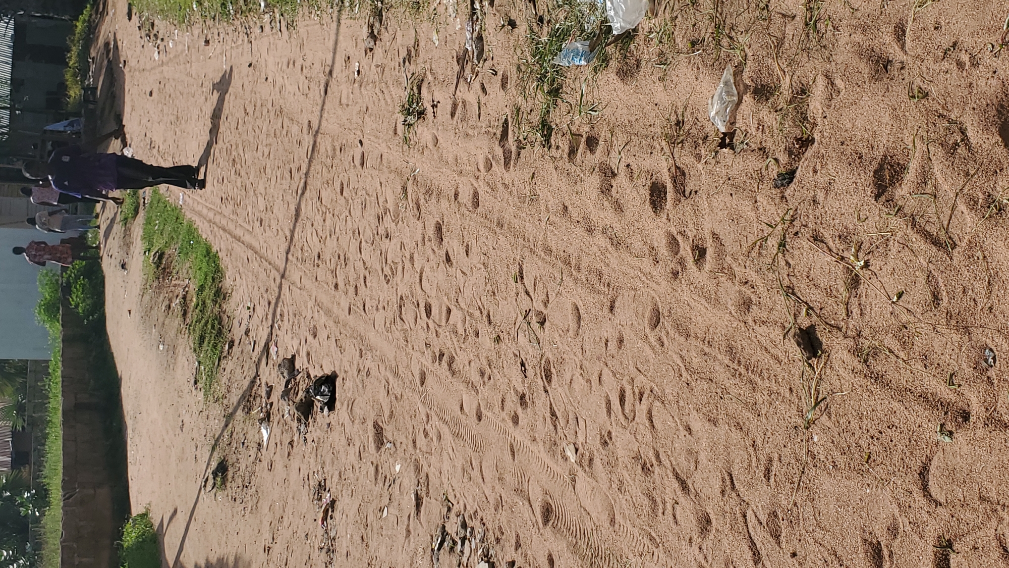 The image depicts a sandy road in Jalingo, Nigeria. The road is marked with tire tracks and numerous footprints, indicating recent activity. Several people are visible in the background, walking away from the camera. The environment appears somewhat neglected, with scattered debris and dry vegetation along the sides of the road. The lighting suggests it is daytime, with shadows cast on the ground.