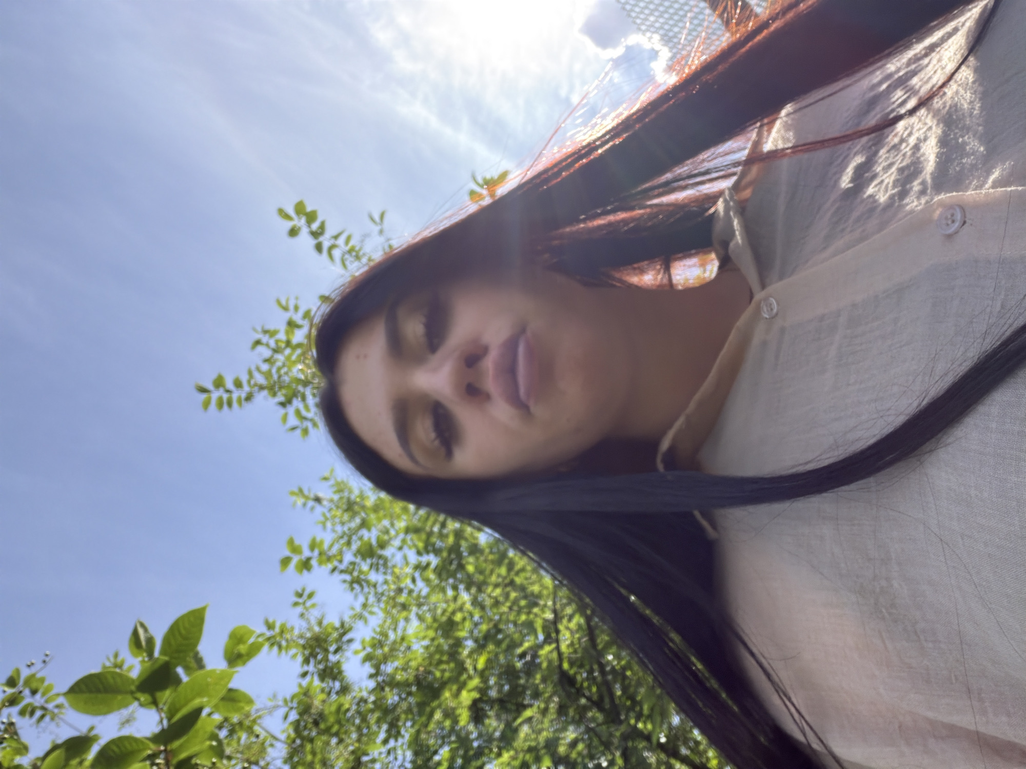 A low-angle, close-up shot of a young woman with long, dark hair, wearing a light-colored collared shirt. She is looking upwards towards the camera with her lips pursed. Green foliage and a bright blue sky with wispy clouds are visible in the background. The image appears to be taken outdoors during daylight, with sunlight glinting off her hair and the right side of her shirt. The setting suggests a natural environment, possibly a park or garden, with no specific identifiable urban features of Фоки, Russia.