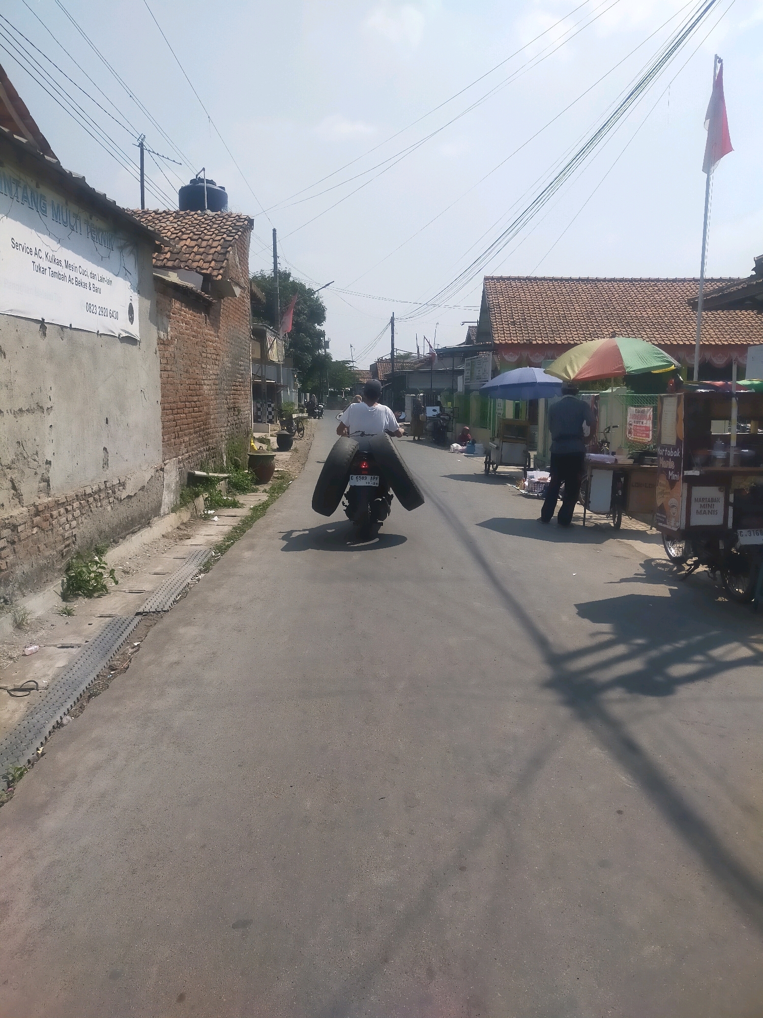 This is a medium shot, eye-level photograph taken on a sunny day on a paved street in Kabunan, Indonesia. The street appears to be a residential or commercial area with single-story buildings lining both sides. In the foreground, a man wearing a white t-shirt and dark pants rides a motorcycle with two large tires strapped to the back. The tires are black and appear to be truck tires. The motorcycle has a license plate visible, though the characters are unclear. The man is riding away from the camera towards the center of the image.
On the left side of the street, a brick building with a rough, gray plastered facade stands. A sign on the building reads "BINTANG MULTI TEKNIK" and lists services such as "Service AC, Kulkas, Mesin Cuci, dan Lain-lain" and "Tukar Tambah Ac Bekas & Baru," along with a phone number. A water tank is visible on the roof of a tiled structure behind this building.
On the right side of the street, there are several street vendors with food carts. One cart has a colorful umbrella and appears to be selling food. Another cart has a sign that says "MARTABAK MINI MANIS." People are visible near the food carts, including a man in dark clothing who appears to be a vendor. A red and white flag is flying from a flagpole on the right side of the street in the background. Power lines crisscross the sky above the street. The overall impression is a bustling, everyday scene in a semi-urban Indonesian environment.