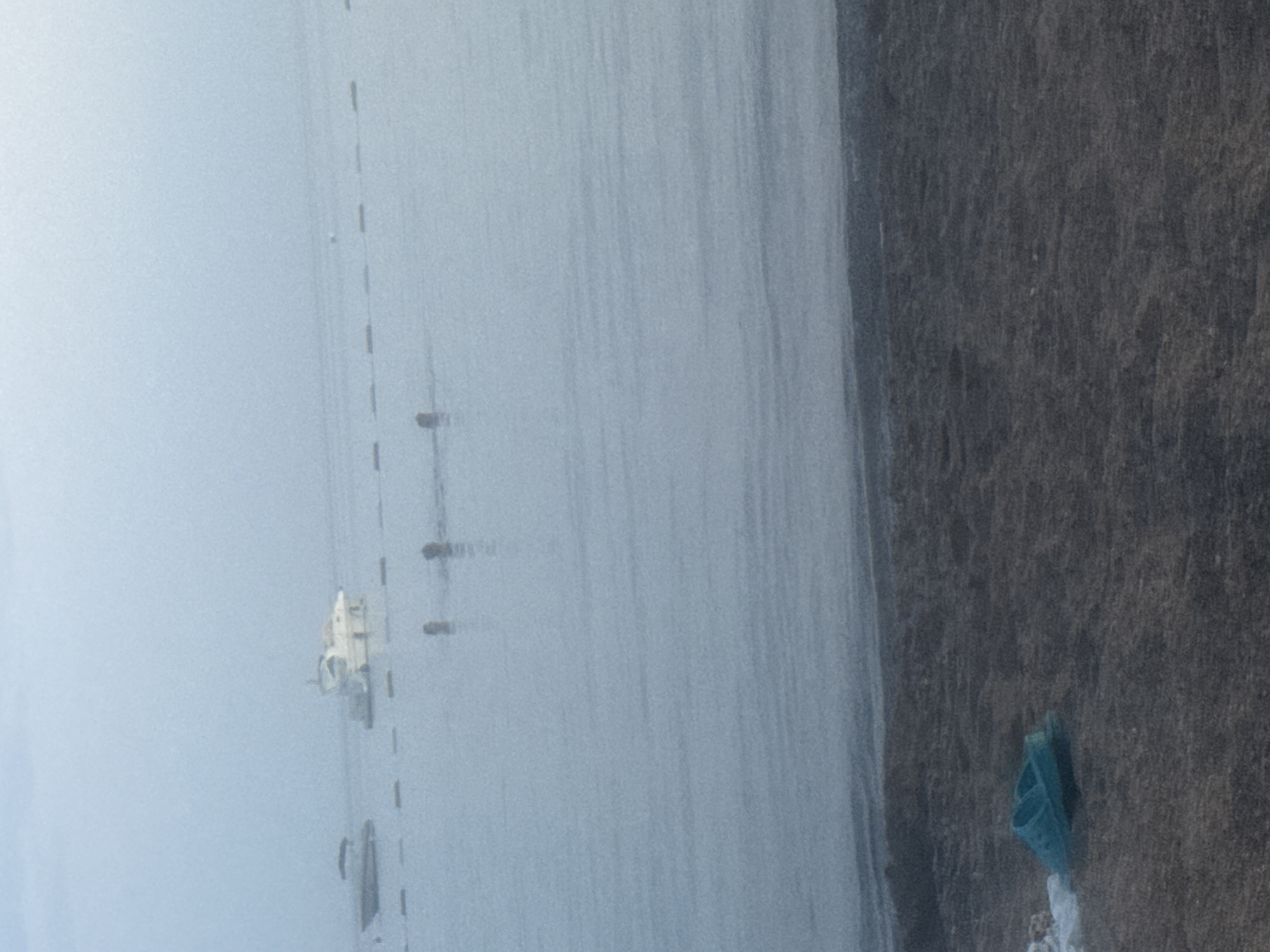 The image shows a slightly hazy view of a calm body of water, likely the Adriatic Sea, near the coast of Rafailovići, Montenegro. In the middle ground, two boats are visible; a smaller, lighter-colored boat on the left and a larger, white yacht with multiple decks on the right. A series of dark, submerged buoys or markers creates a dotted line across the water. In the water, several human figures are partially submerged, appearing to be swimming or wading. The foreground depicts a dark, sandy beach with a pair of bright blue flip-flops lying near the water's edge. The overall atmosphere is serene and possibly early morning or late evening due to the soft light and haze.