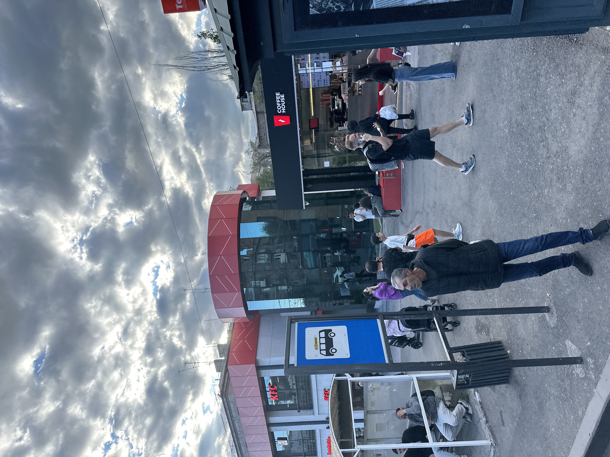 The image shows a street scene in Yerevan, Armenia, identified by the presence of a "Coffee House" cafe and a KFC restaurant. People are walking and waiting near a bus stop, indicated by a blue sign with a bus symbol. The sky is overcast with dramatic cloud formations. Several people are visible, including individuals in casual clothing. The architectural styles of the buildings suggest an urban environment.