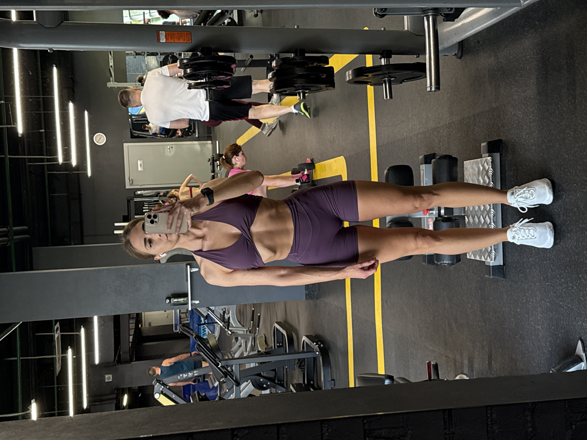 A woman with brown hair pulled back and wearing a purple athletic set is taking a mirror selfie in a gym. She is wearing white sneakers and wireless earbuds. In the background, other gym-goers are visible, including a man in a white t-shirt and black shorts lifting weights. The gym appears to be located in Saint Petersburg, Russia, as indicated by the presence of Cyrillic text on a sign in the background that reads "РАБОТАЕТ" (works) and the overall aesthetic of the facility. The gym has industrial-style lighting, dark grey walls, and a black rubberized floor with yellow safety lines. Weightlifting equipment, including weight racks and machines, are visible throughout the space.
