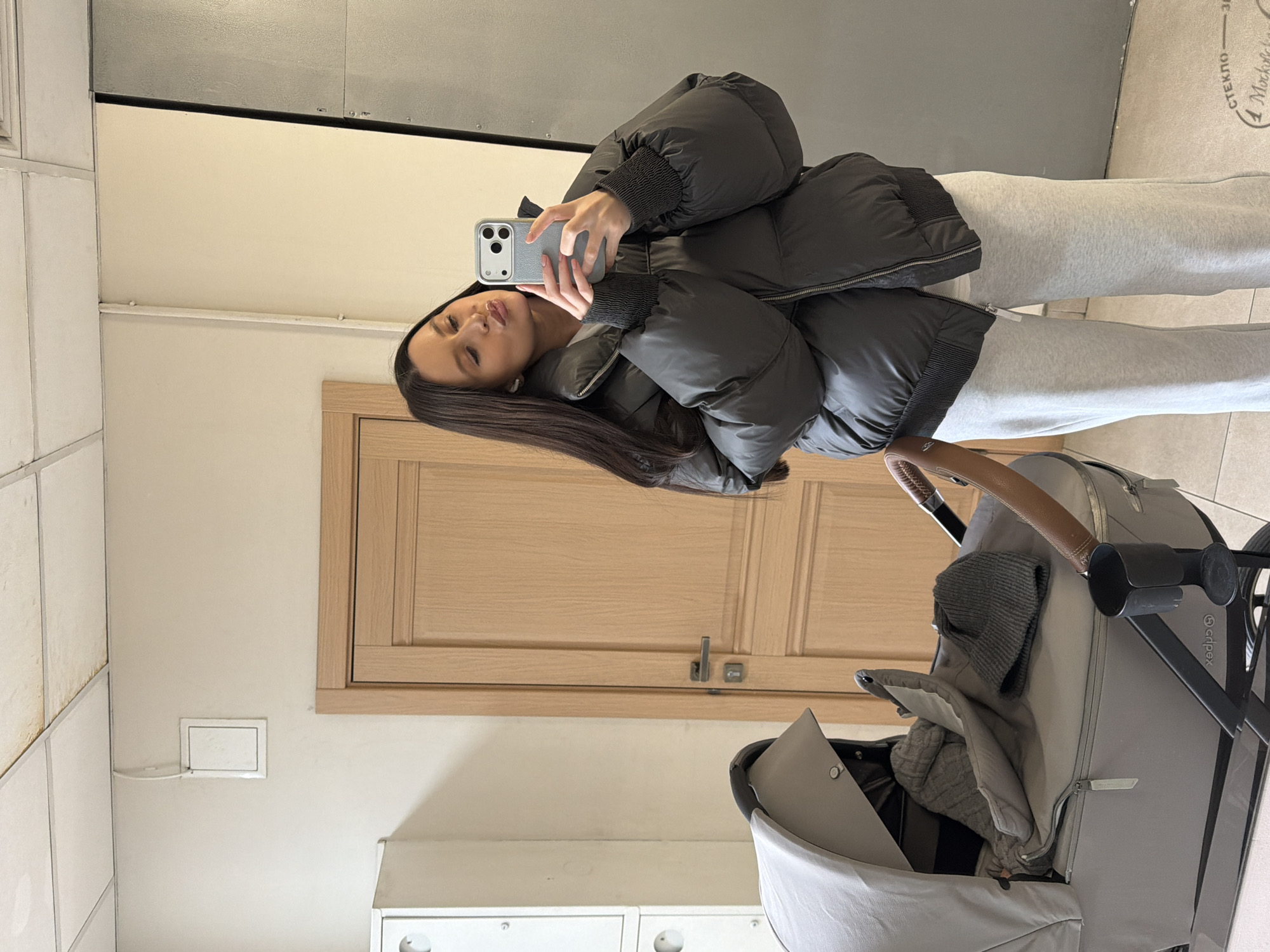 The vertical image, rotated counter-clockwise, depicts a woman taking a mirror selfie indoors. The woman has long dark hair and is wearing a dark grey or black puffer jacket with ribbed cuffs and light grey sweatpants. She holds a grey smartphone, likely an iPhone, up to the mirror to capture her reflection. She has a posed expression with visible lip gloss.
To her right, a modern grey baby stroller with a brown leather-look handle and a black frame is parked. A grey knitted hat and a blanket are visible inside the stroller, with the brand "Cybex" partially visible on its frame.
The background shows an indoor space, possibly a hallway or common area. The walls consist of white tiled panels on the left, a plain cream-colored section, and a light wooden door with a handle. A light wooden cabinet is visible above the door. The ceiling includes a dark grey or metallic panel section. A white electrical outlet or access panel is mounted on the cream wall.
Reflected in the upper right corner of the mirror, Russian text is visible, including "СТЕКЛО" (meaning "Glass") and "Москве" (meaning "in Moscow" or "Moscow's"). The puffer jacket suggests cooler weather, aligning with the given location context of Khimki, Russia.
