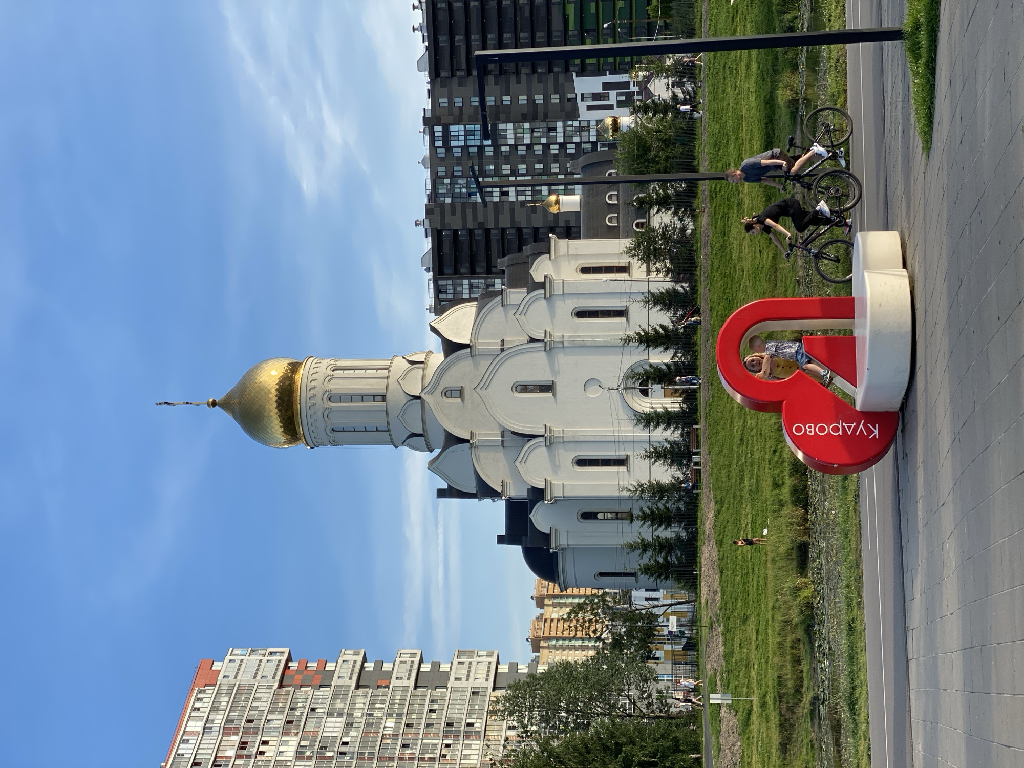 In the city of Dnp Ryzhiki, Russia, a white orthodox church with a golden dome stands prominently against a clear blue sky. The church features a tall, octagonal bell tower with arched windows and decorative cornices. Adjacent to the church, modern residential buildings with multiple floors and balconies are visible. In the foreground, a large red heart-shaped sculpture is positioned near a paved walkway and a road. Two individuals are cycling on the road, while a child sits inside the heart sculpture. A grassy area with trees lies between the walkway and the church.