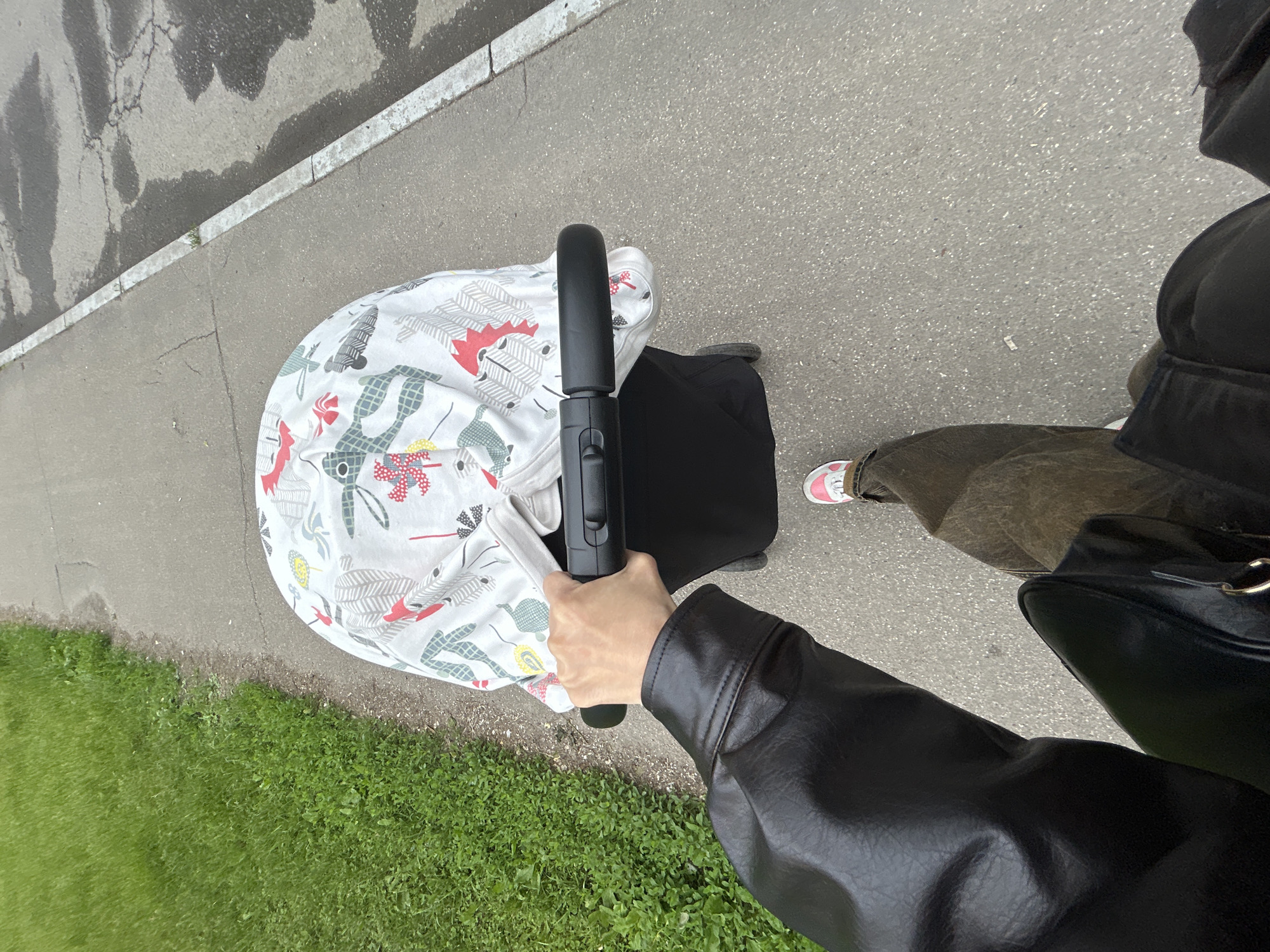 The image is a first-person perspective shot, looking down as someone pushes a stroller along an outdoor path. A person's right hand and forearm, covered by a black faux-leather jacket, firmly grasp the black handle of a stroller. The stroller itself features a white fabric cover with colorful, stylized patterns that include animal-like figures and geometric shapes in red, gray, blue, and yellow. To the left of the path, a vibrant strip of green grass is visible. On the right, the person's lower leg, clad in dark trousers and a white sneaker with a pink detail (possibly a logo), is seen. The ground beneath the stroller and the person is an asphalt pathway, which borders a cracked, darker asphalt road or shoulder, separated by a distinct white painted line. The lighting is bright and even, indicating daytime with diffused sunlight. No specific text is visible in the frame. The scene captures a casual walk outdoors, likely in an urban or suburban setting.